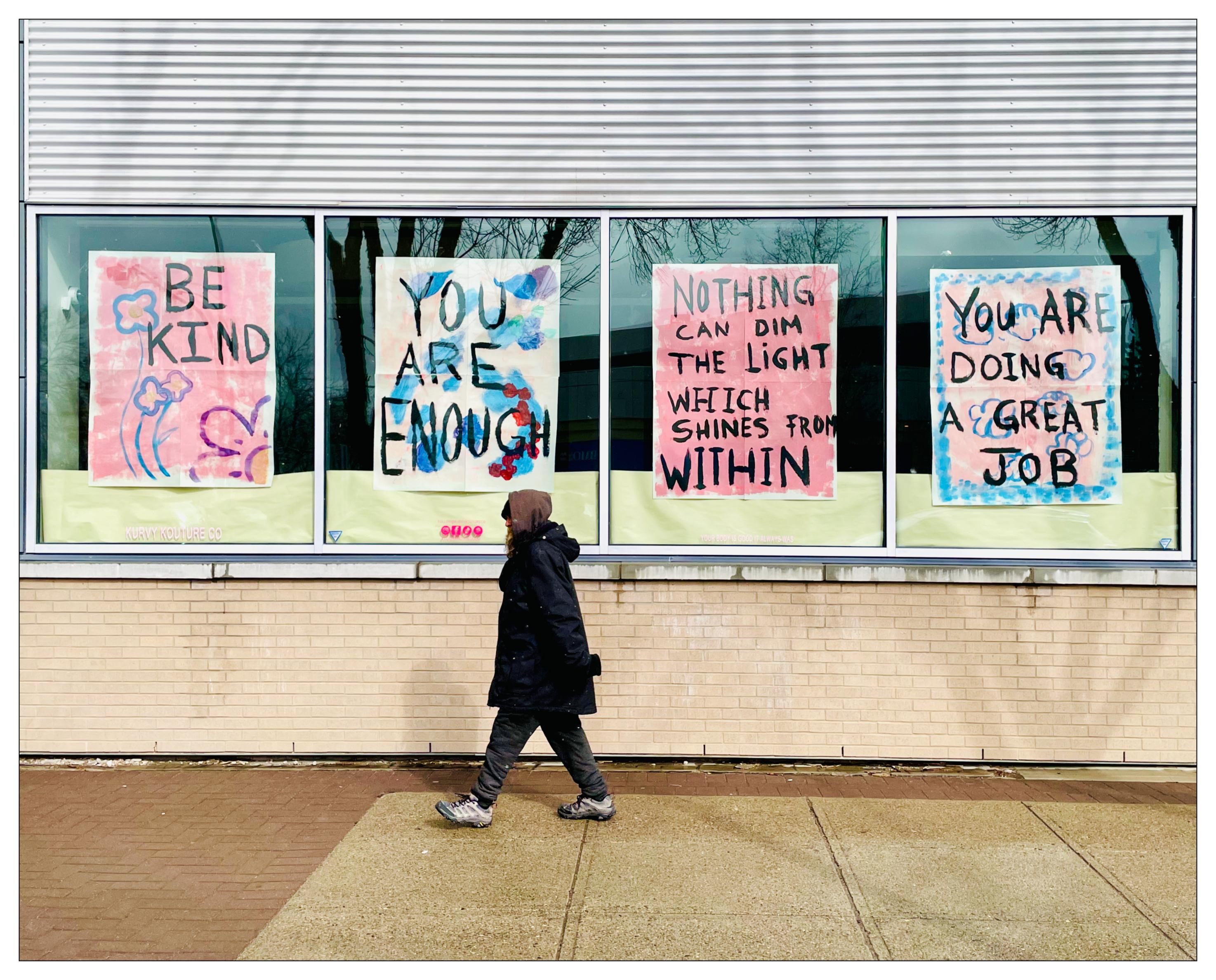 Person in dark clothes walking past a vacant store front with high windows. Posters in the windows, hand painted, say
Be Kind
You Are Enough
Nothing can dim the light which shines from within
You are doing a great job