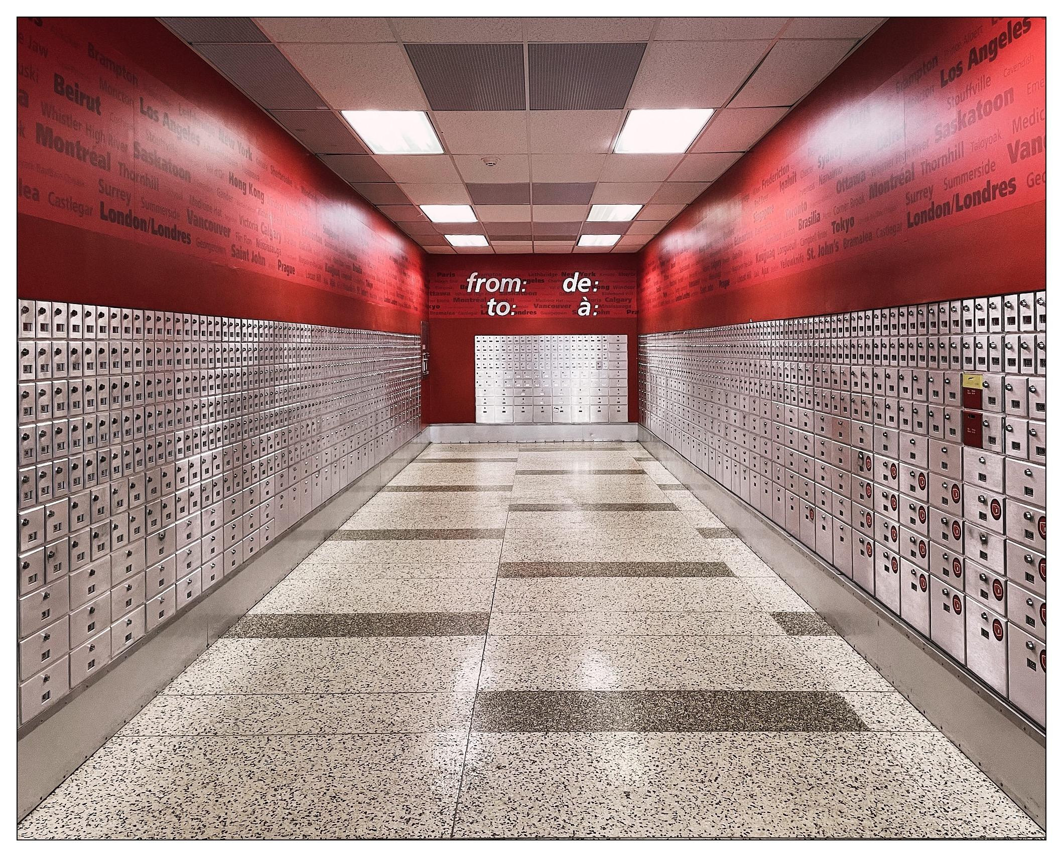 Interior of a mailroom with bright red walls and rows of white and gray metal postal boxes. The walls are covered with lists of city names and directional phrases like "from:" and "to:" in various sizes and bold lettering. The floor is a patterned mix of light gray, dark gray, and tan tiles. The ceiling features flush lighting, providing even illumination throughout the space.