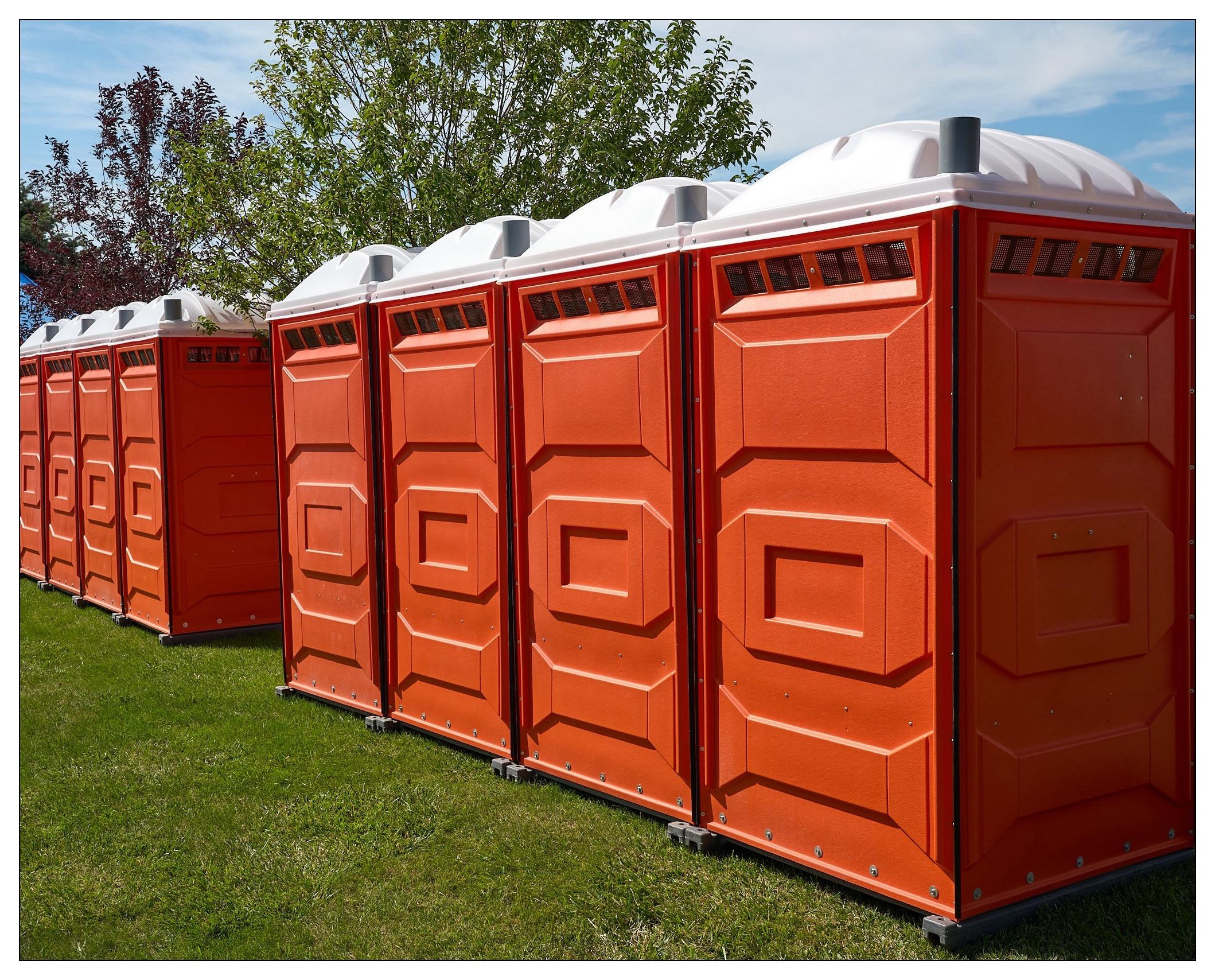 Row of bright orange portable toilets lined up on grass, with the endmost units partially visible. Each toilet has a white roof and geometric patterns on the doors, set against a background of trees and a blue sky with light clouds.