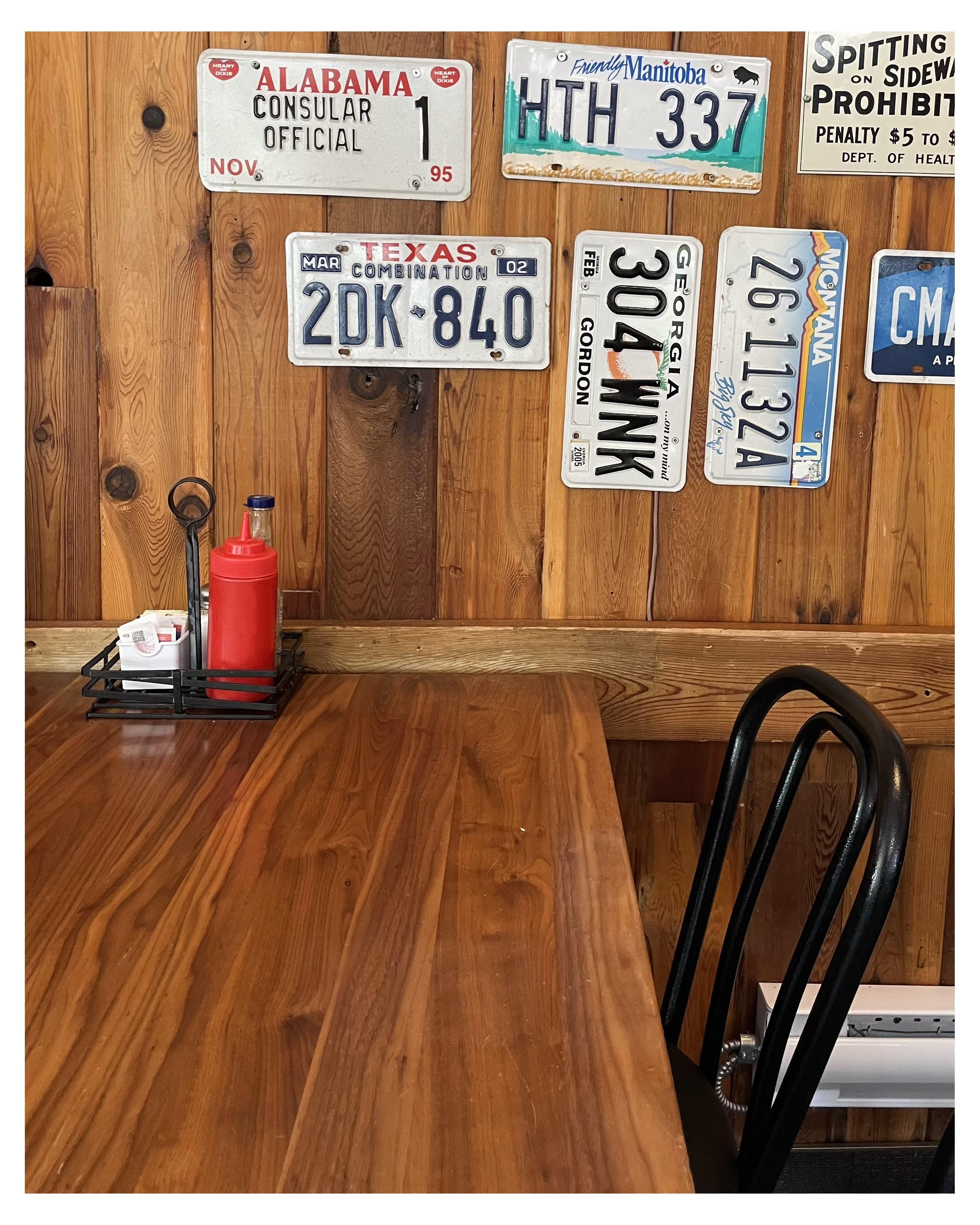 Wooden diner table with metal chairs in the foreground. Wall behind is lined with various international and American license plates including ones from Alabama, Texas, Georgia, and Manitoba. A condiment caddy containing ketchup and menu cards sits on the table.