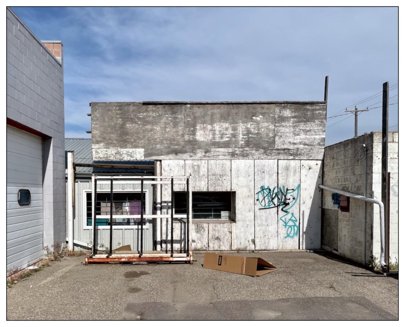 Weathered building with a flat roof, featuring a patched-up facade of white and gray panels. The front of the building includes a set of metal-framed glass doors beneath a faded sign, barely visible. To the right, graffiti in blue and green adorns the wall. A fallen cardboard box lies on the concrete foreground, and adjacent structures partially enclose the scene under a clear sky.