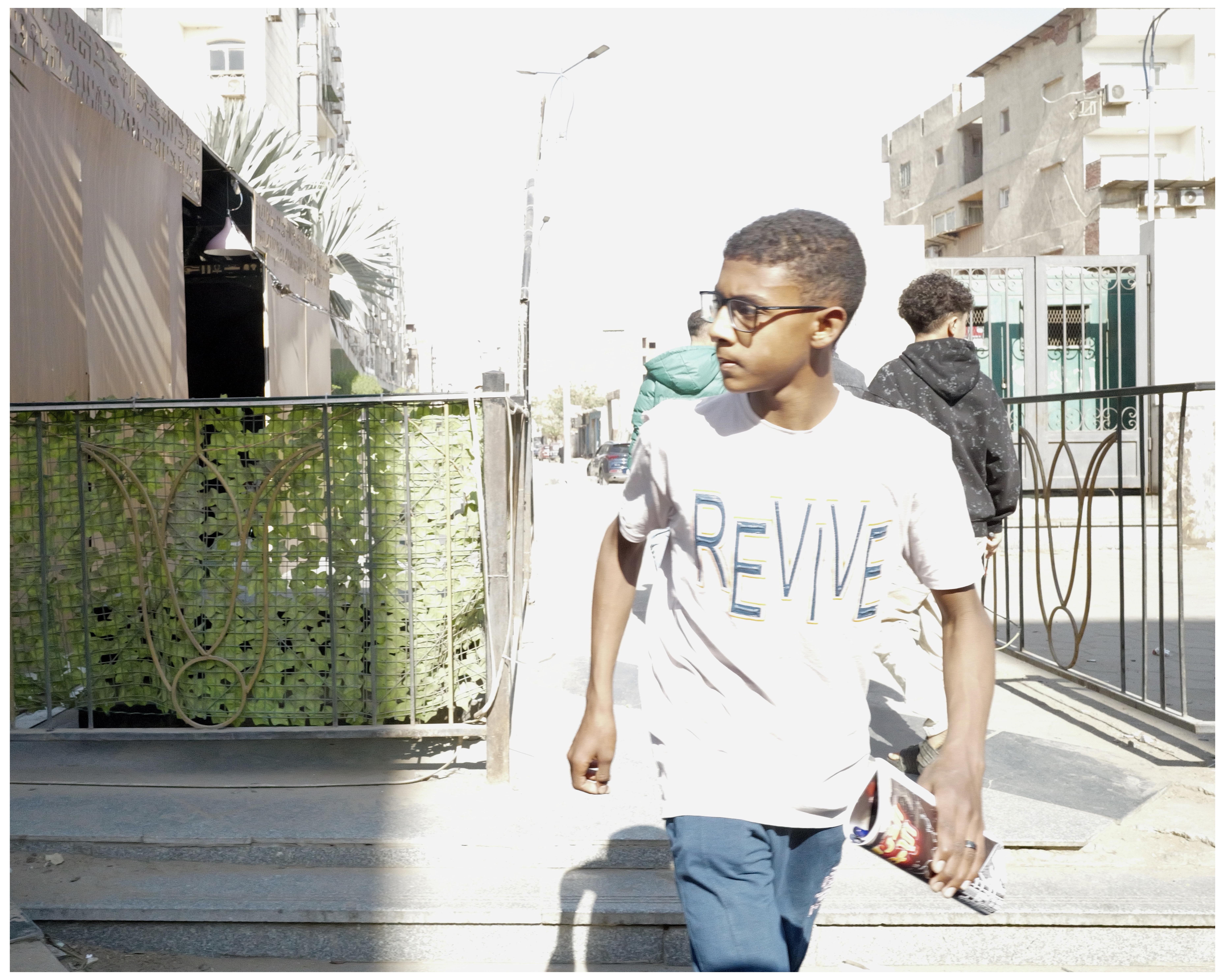 In the image, there is a young male wearing glasses and a white t-shirt with the word "REVIVE" printed on it. He is walking on a sidewalk holding what appears to be a brochure or a booklet. The setting seems to be a city street, with buildings and a metal gate partly visible. A couple of other people can be seen in the background. The overall atmosphere looks quite sunny and bright.