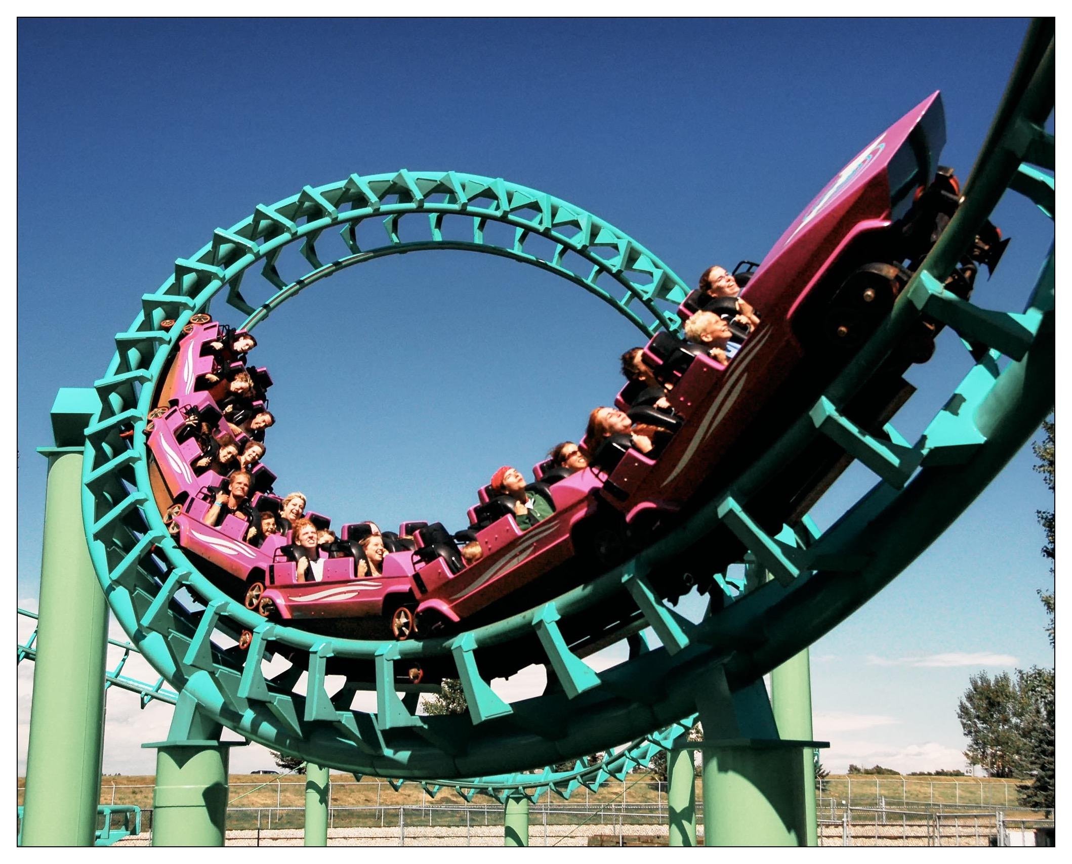 This image shows a group of people enjoying a ride on a roller coaster, which is depicted at the moment it is navigating through a loop. The roller coaster has a light green track and is supported by a turquoise-colored structure. The sky is clear and blue, indicating nice weather, which adds to the vibrancy of the scene. The passengers appear to be experiencing excitement and thrill, typical of such high-adrenaline amusement park rides.