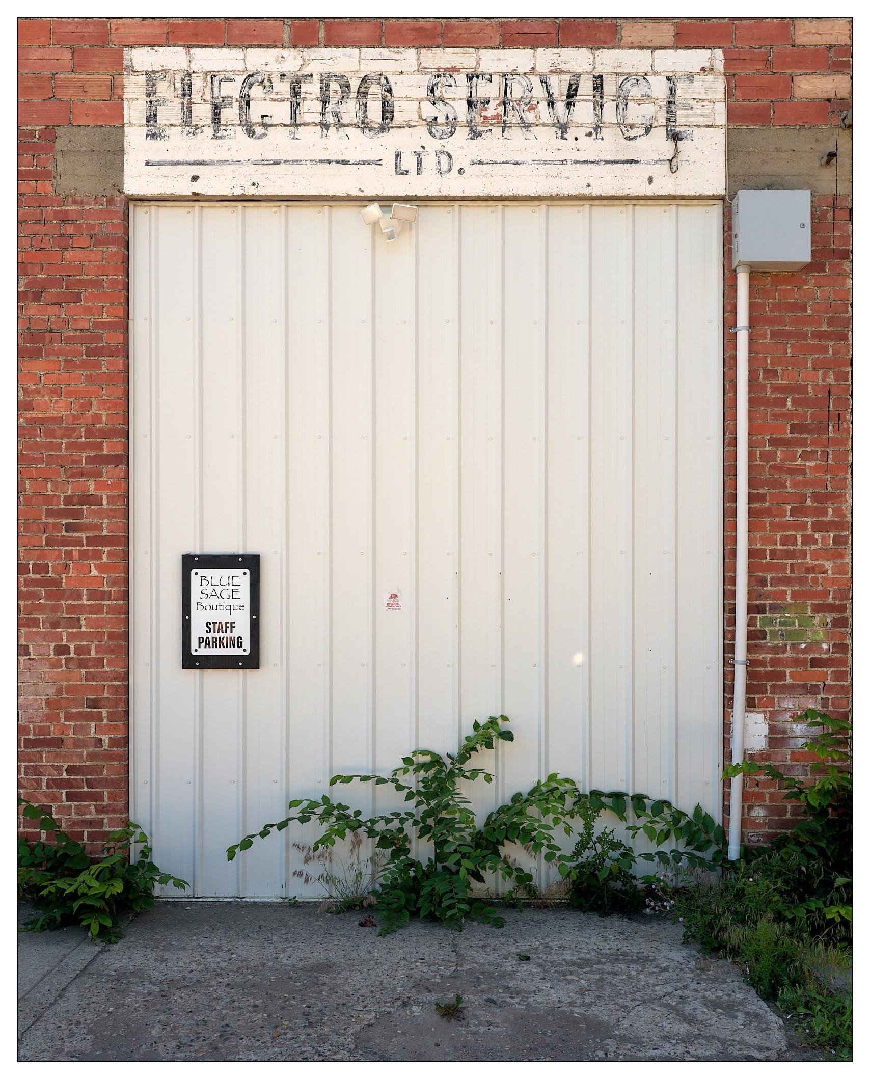 A white garage door set into a brick building, with "ELECTRO SERVICE LTD." painted in black on a fading white sign above. To the right of the door, a small rectangular sign reads "BLUE SAGE BOUTIQUE STAFF PARKING." Weeds and green plants grow along the base of the wall and crack-filled concrete ground.