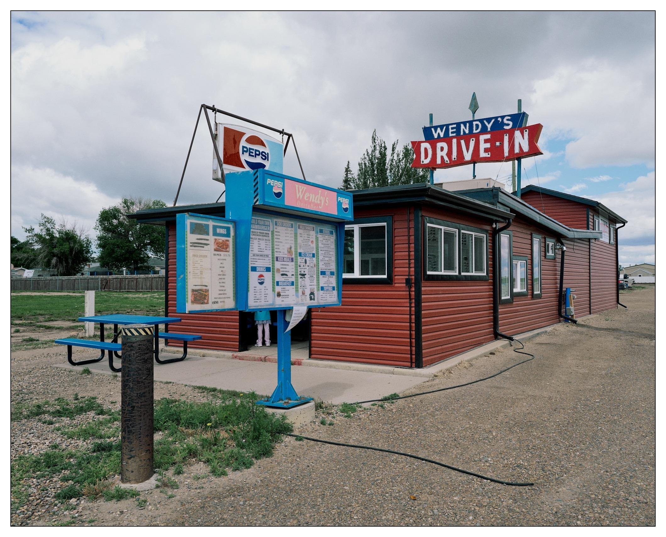 Small town drive-through fast food.  Sign says "Wendy's Drive-In" but it's not related to the big chain of the same name.  There are pneumatic hoses to ring a bell when a vehicle pulls up to order,  a price sign board, a faded glass light up Pepsi sign, and a newer metal blue and black picnic table visible.