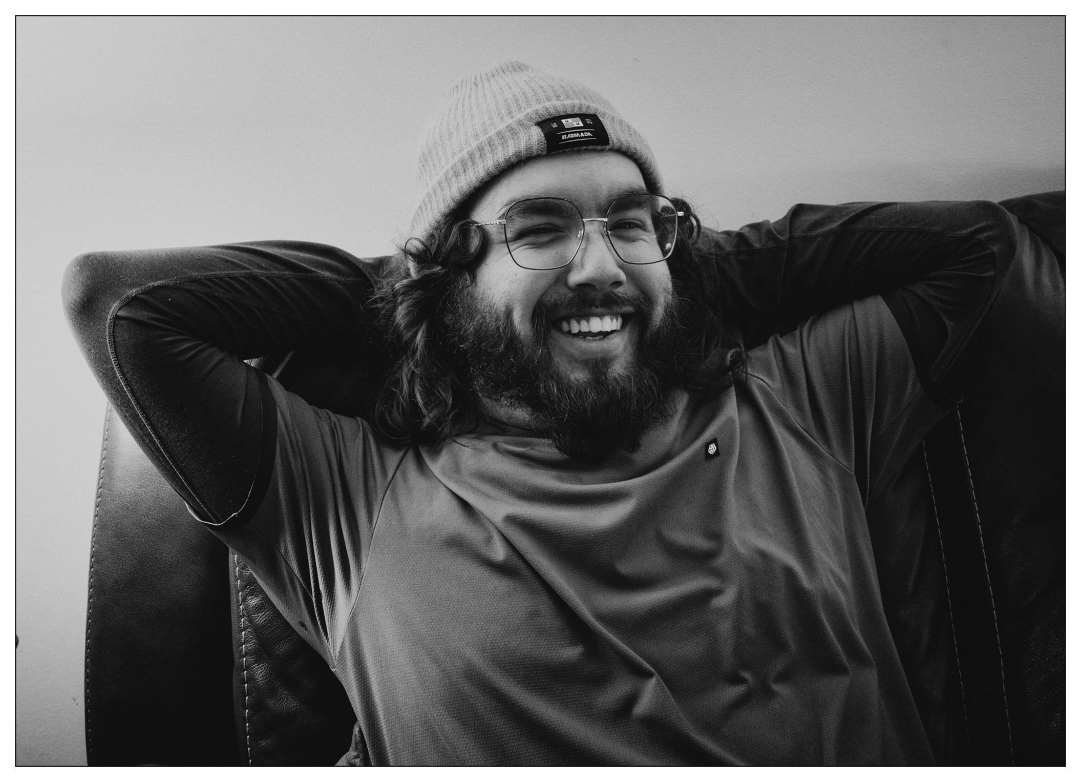The image shows a cheerful man with long, curly hair wearing glasses, a beanie hat, and a turtleneck top. He is sitting in what appears to be a high-backed leather chair, laughing or smiling broadly and resting his hand behind his head. The photo is in black and white, which adds a classic, timeless feel to the shot. The man exudes a relaxed and joyful demeanor.