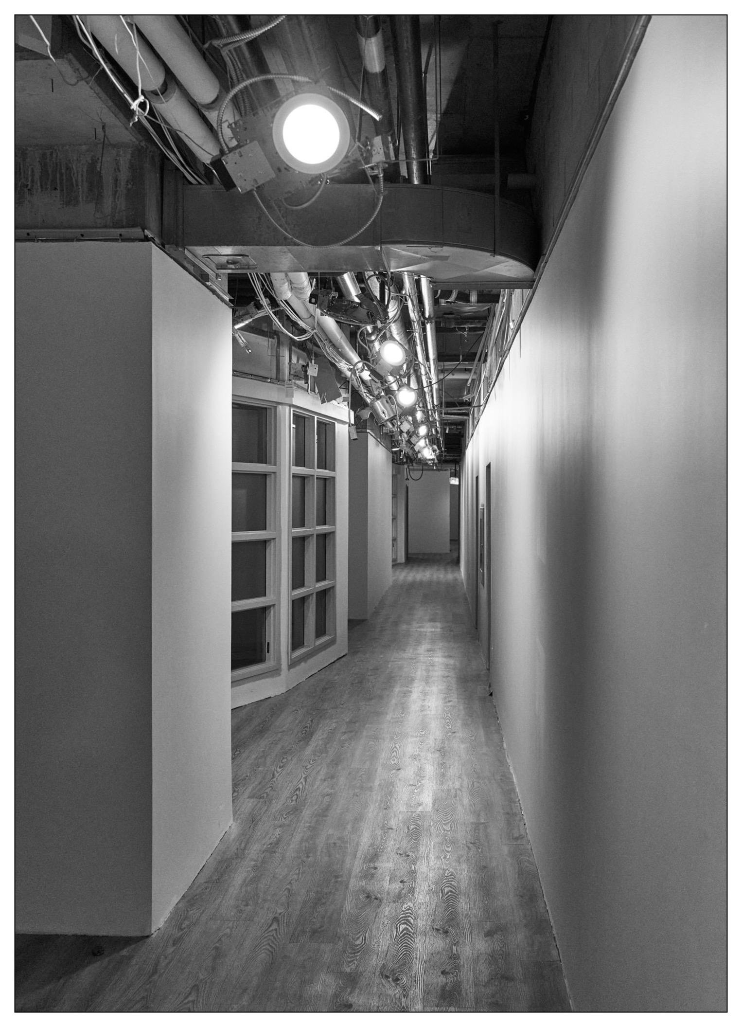 Black and white photograph of a long, narrow corridor in an industrial or office setting with exposed overhead ductwork and lighting. The left side of the corridor features a row of closed windows with white frames, while the right side has a plain wall. The floor is covered with light wooden planks, and multiple spotlights are mounted along the ceiling, casting bright spots of light at intervals along the corridor.