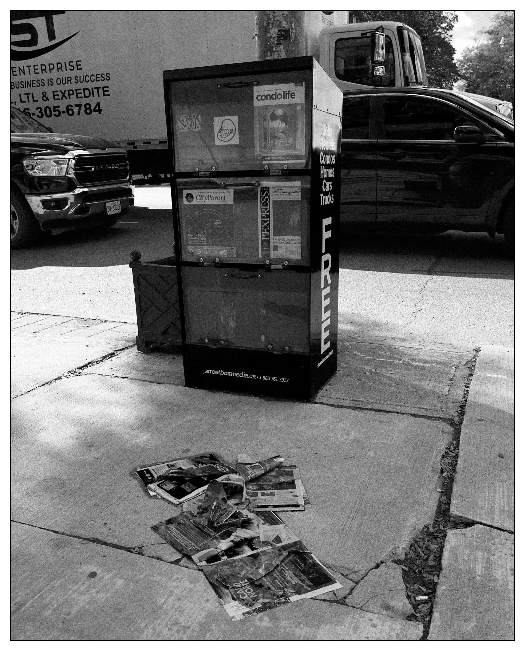 Black and white photograph featuring an urban streetscape with two relevant objects in the foreground: a newspaper vending machine labeled "FREE" with various publications visible, and a pile of discarded papers scattered on the sidewalk. In the background, part of a parked truck with a business advertisement and a parked black SUV are visible. The scene captures elements of city life and discarded items, highlighting urban clutter.