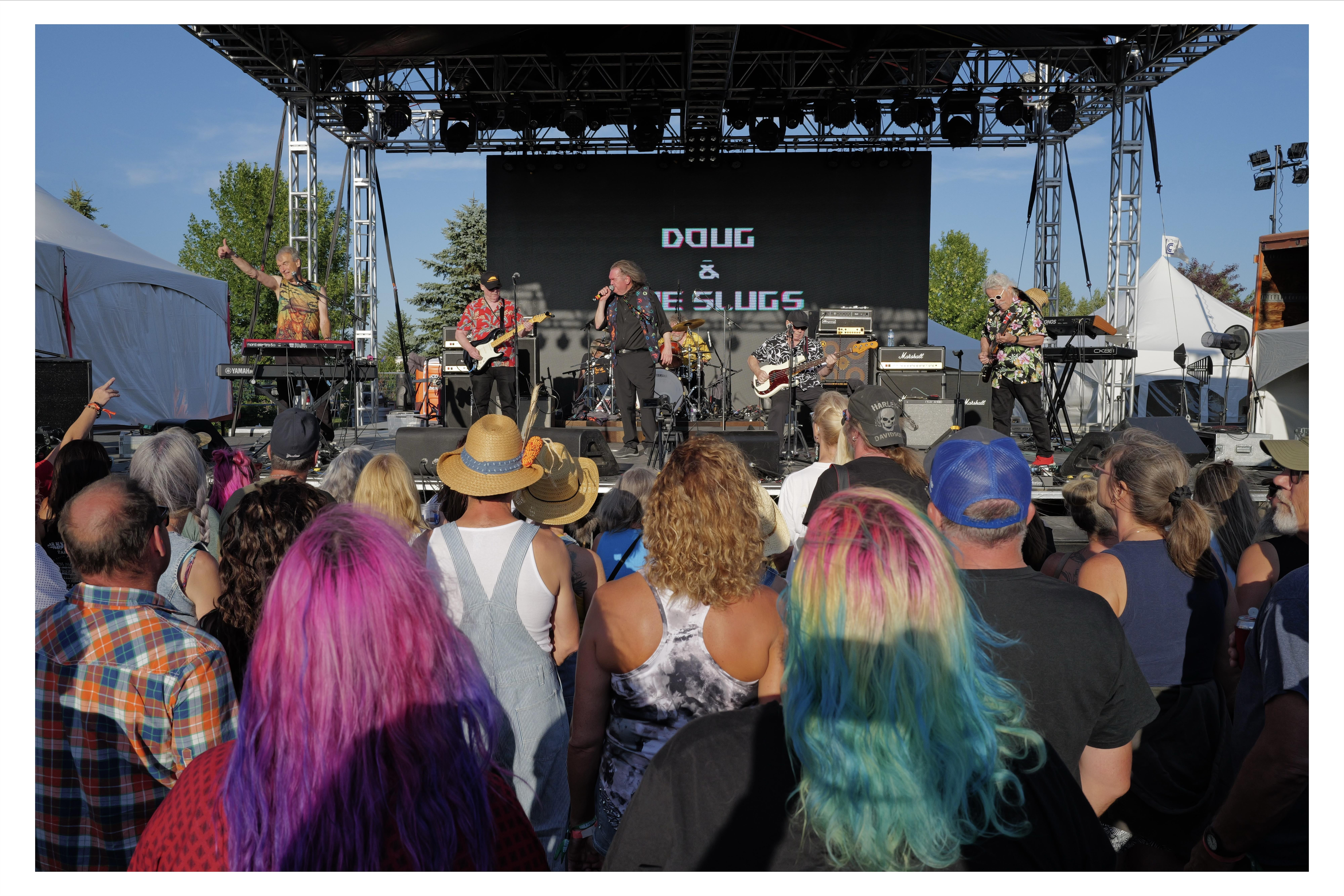 Outdoor music concert with a crowd watching the band 'Doug & The Slugs' performing on stage. Several band members are visible; some playing guitars and another on keyboards. The audience in the foreground includes people with brightly colored hair, facing the stage, some with raised hands. It's a sunny day with clear skies and trees in the background.