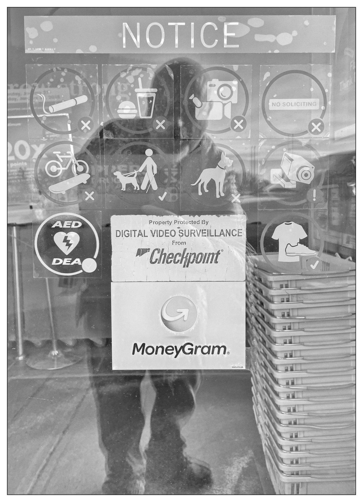 The image shows a glass door or window covered with various notices and symbols. At the top, there is a large "NOTICE" label indicating that the premises are digitally monitored by video surveillance, denoted by the Checkpoint logo below. Various prohibited actions are displayed in circular symbols, such as no cameras, no drinks, no soliciting, no skateboarding, no bikes, no dogs, among others. Additionally, there is a label revealing the presence of an Automated External Defibrillator (AED) on the premises. Below, there is a promotional sticker for "MoneyGram." The background prominently features a reflective surface showing a silhouette of a person taking the photograph.