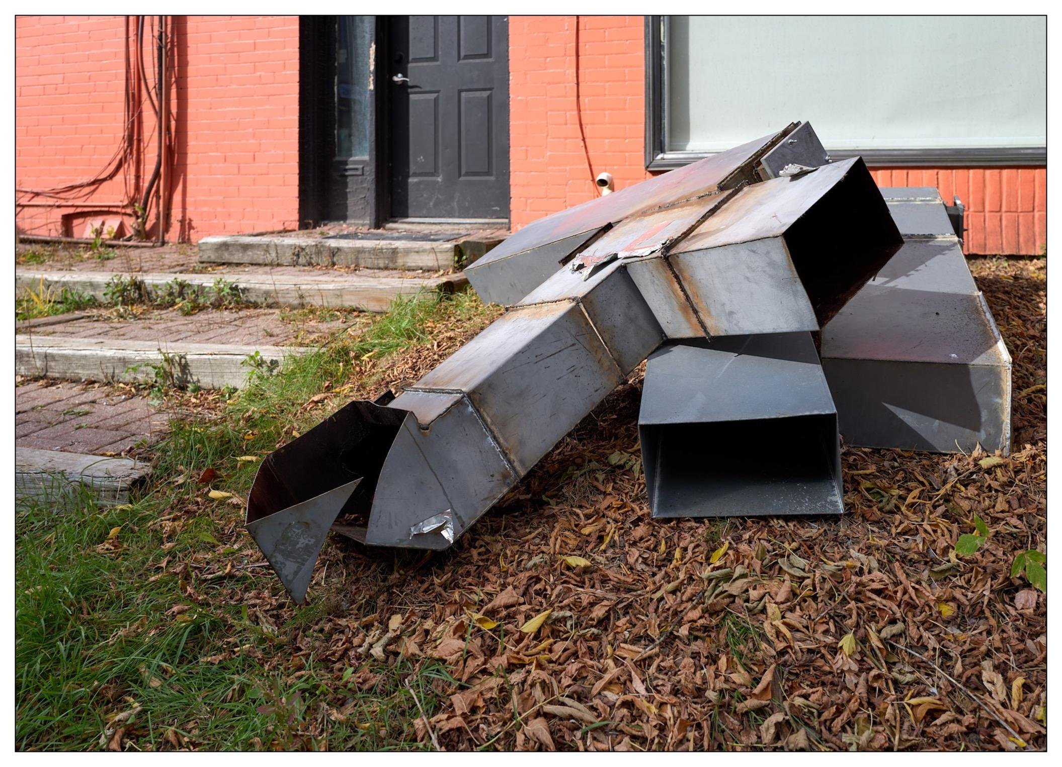 Several large, disjointed metal duct pieces scattered on a bed of dried leaves in front of a red brick building with a black door.