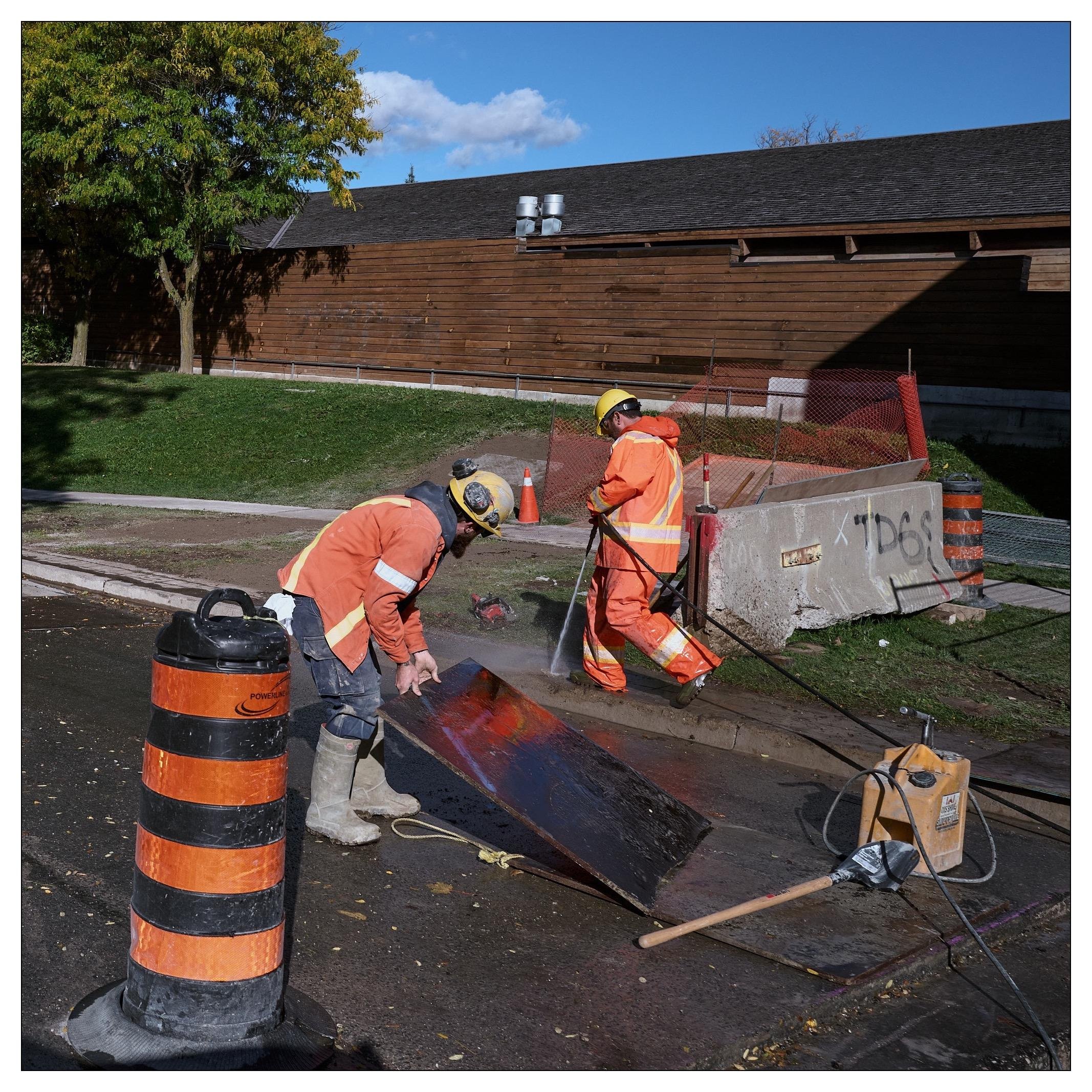 Two construction workers in orange safety gear are working on a road repair. One, wearing a helmet, is lifting a freshly cut piece of asphalt while the other, with a safety harness, uses a hose from a small portable water tank to wash the area. Orange traffic cones and a large, tagged waste container are nearby. Behind them, a wooden building under a blue sky partly obscures the view.