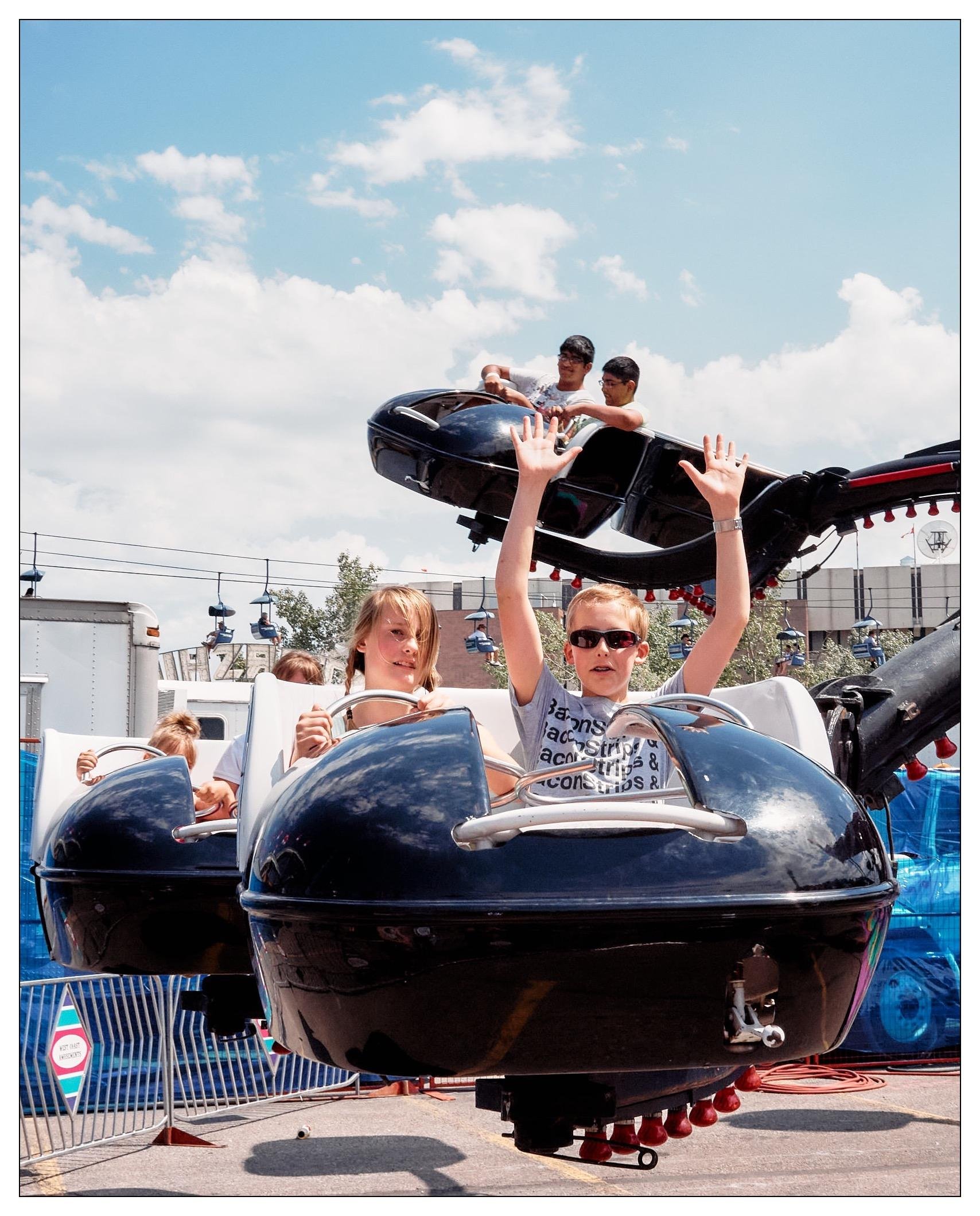 The image depicts a joyful scene at an amusement park or fair where children are riding in a spinning ride, probably the Octopus. There are two kids prominently displayed in the foreground who look exhilarated, with one of them raising their hands high in excitement. The ride itself appears to be designed like a series of small black pods connected to a rotating center, typical of amusement park rides aimed at providing a fun, mildly thrilling experience for kids. The background features a clear blue sky scattered with some clouds and other amusement park installations, enhancing the festive atmosphere of the setting.