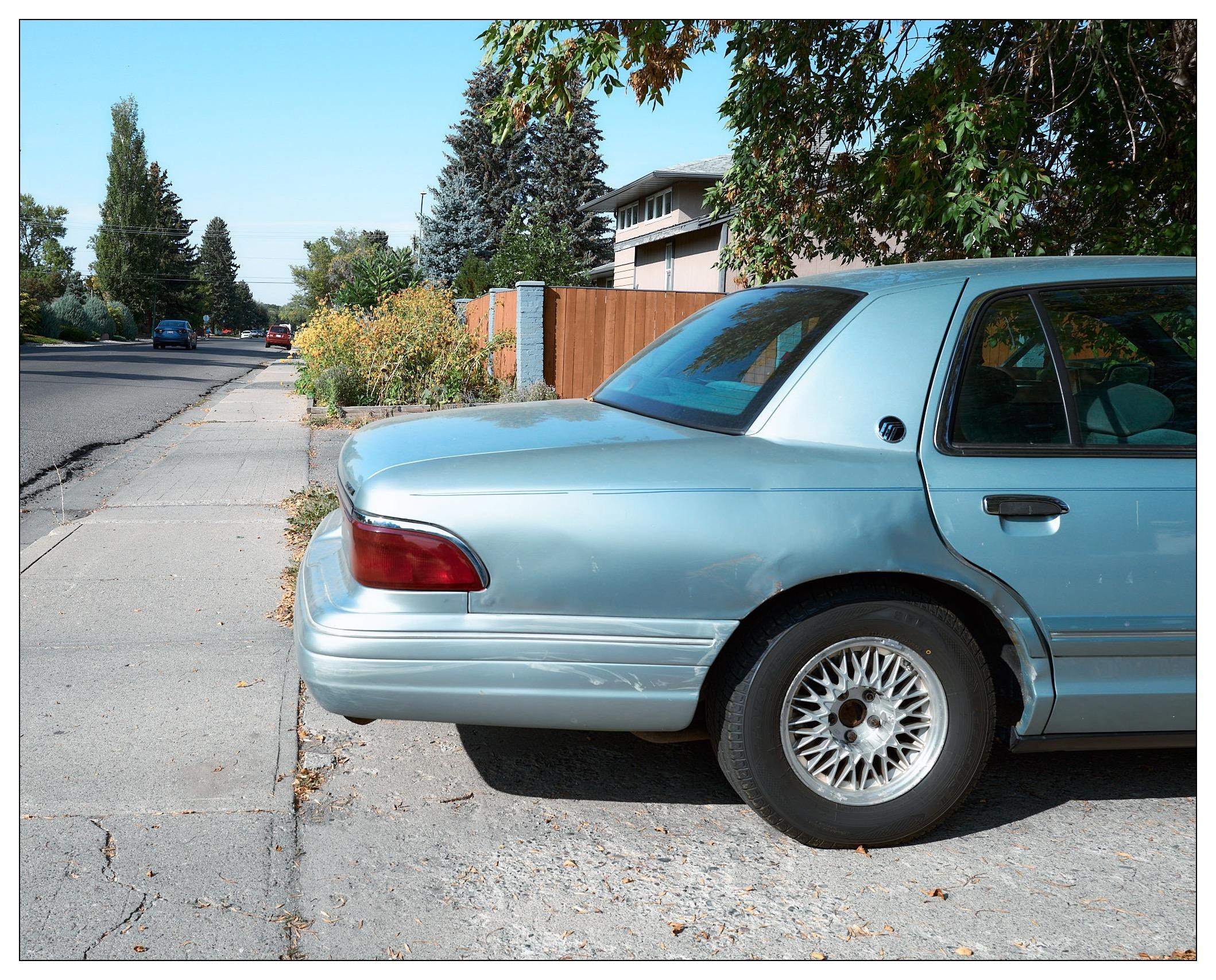 Light blue sedan parked on a suburban street beside a curb, featuring a distinct shiny wheel design. Trees, a wooden fence, and another distant car on the road are visible in the background under a clear blue sky.