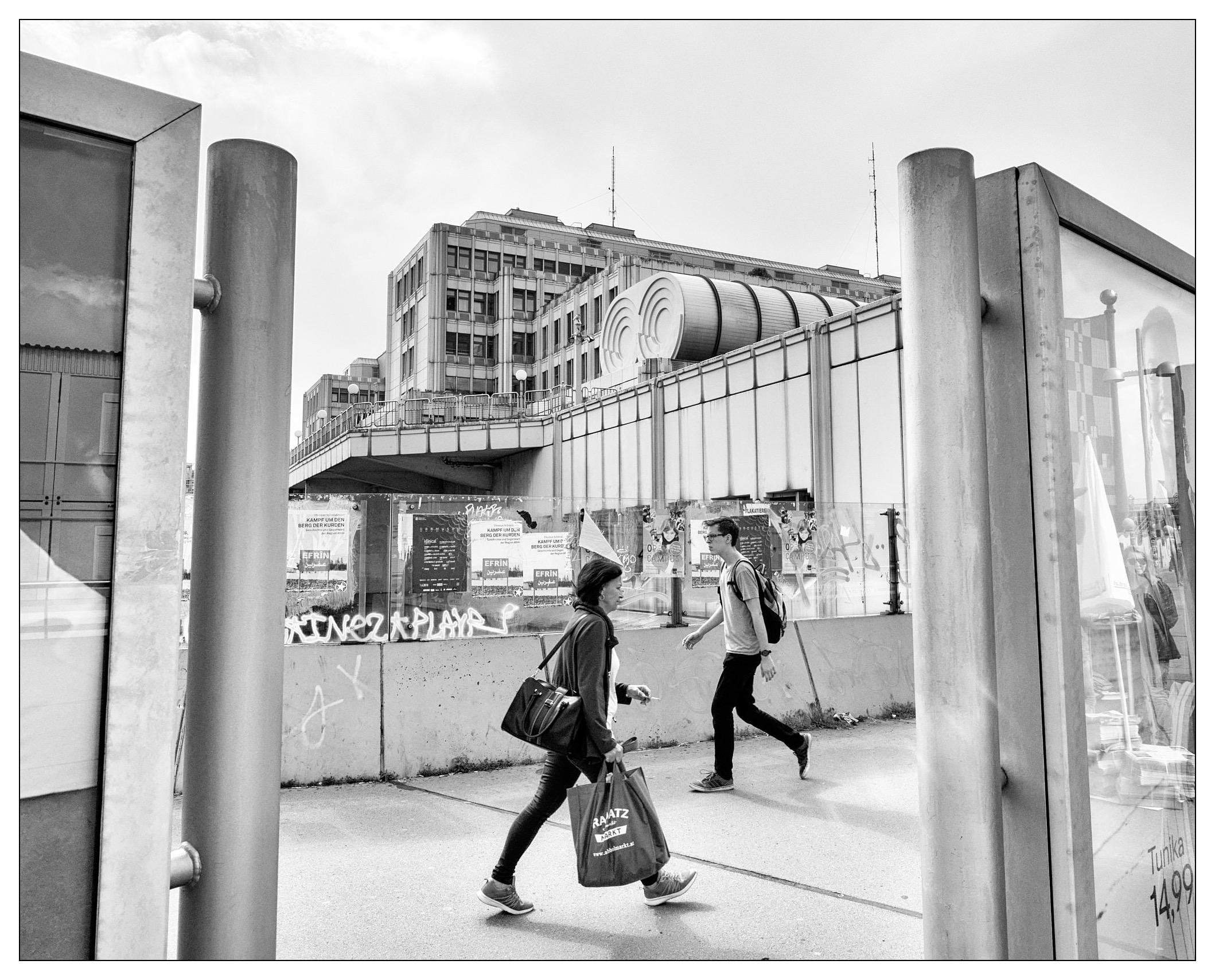 This black-and-white image captures an urban street scene with two pedestrians walking past each other. To the right, there's a large advertisement visible through a glass panel, and in the background, there are industrial or commercial buildings including a distinctive round structure that could be part of a transportation system or a similar public facility. The setting includes urban infrastructure like fencing and graffiti-covered pillars, reflecting a bustling city environment. The image is dynamic, showcasing everyday city life with architectural and street elements.