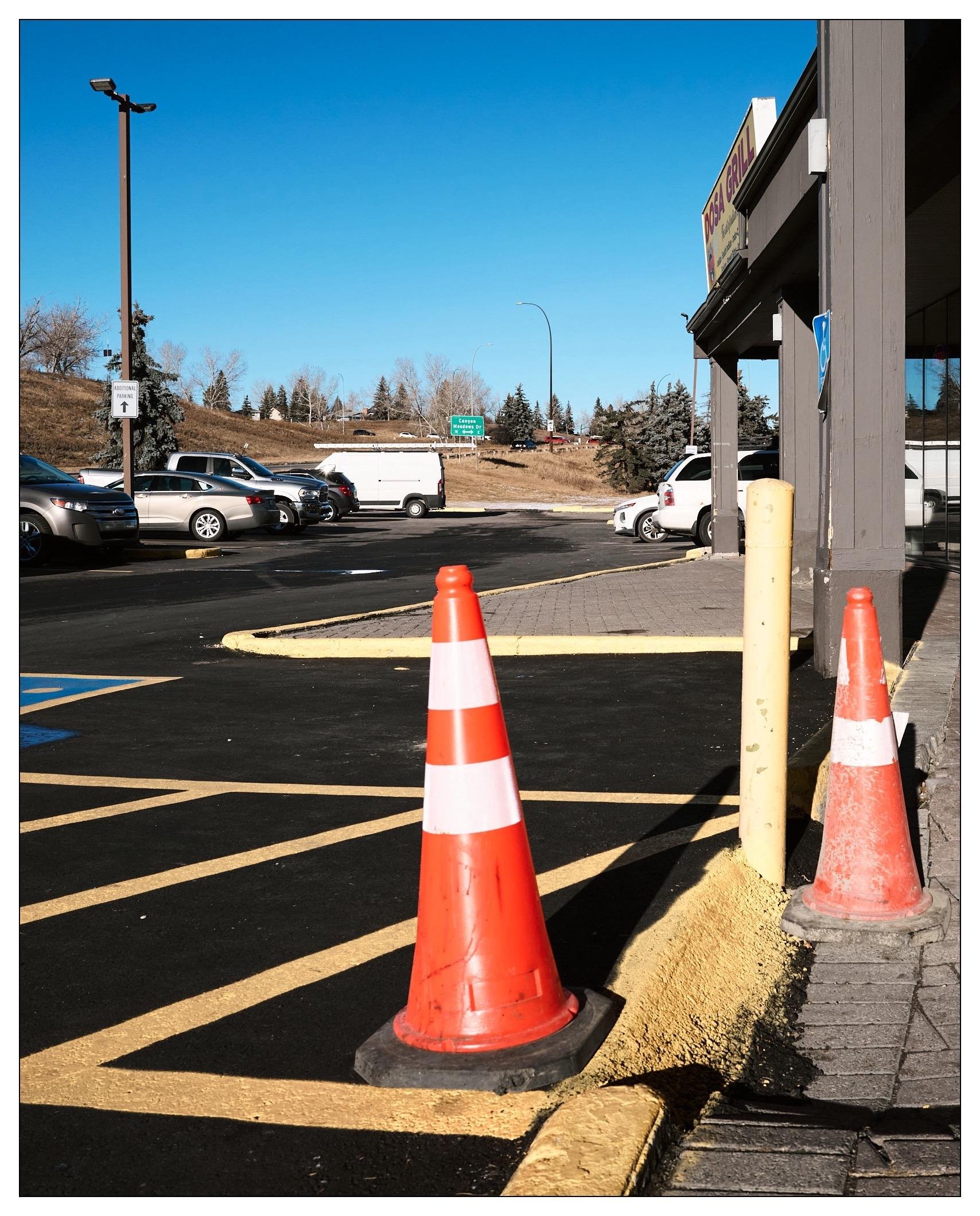 Orange traffic cones on a parking lot with yellow and black striped curbs, adjacent to a building with signage and a parking lot full of vehicles in the background. A lamp post stands on the left near the building, and a clear blue sky is visible overhead.