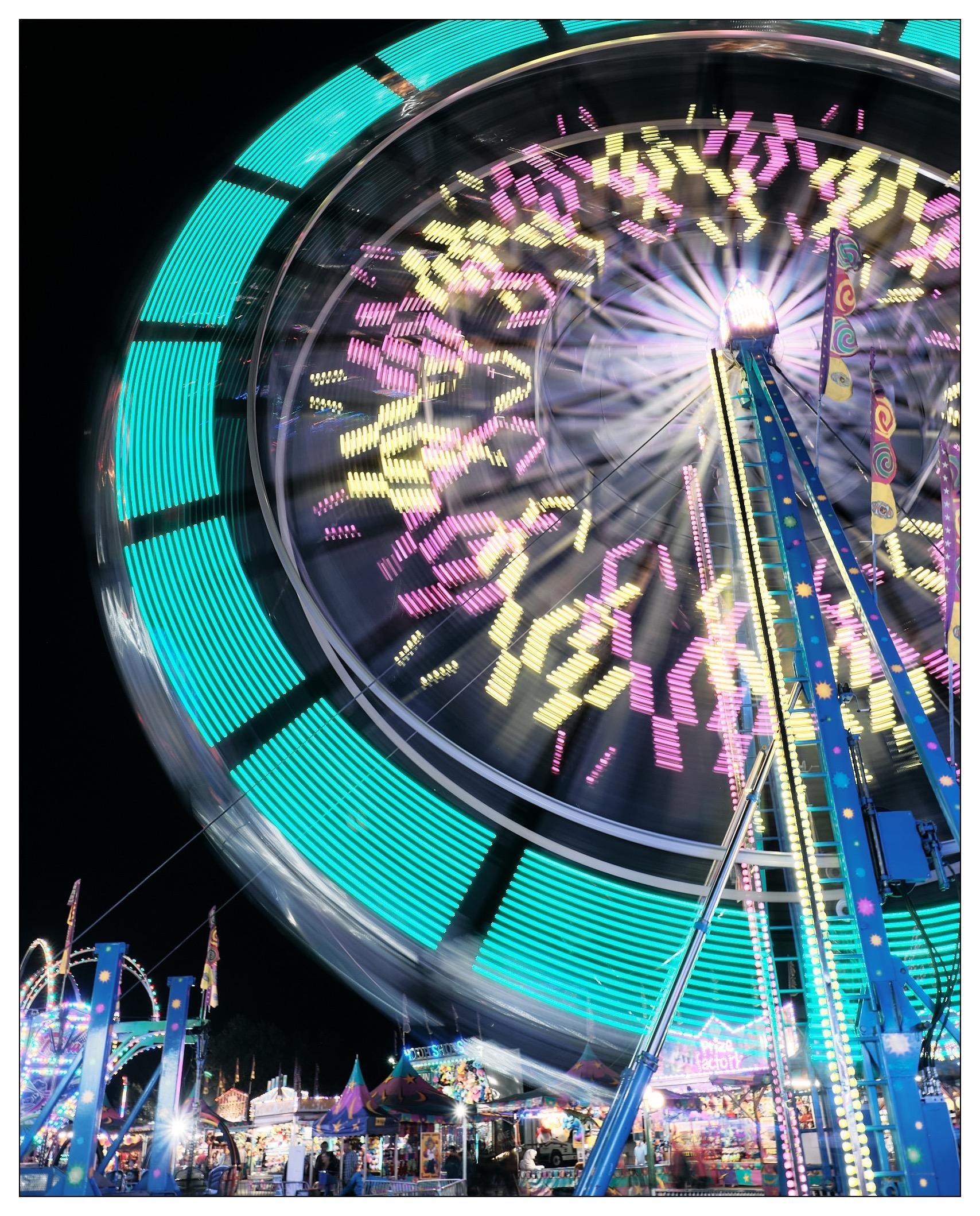 This image captures a vibrant and bustling amusement park at night, featuring a large Ferris wheel in motion. The Ferris wheel is lit with colorful LED lights, and the long exposure used in the photograph allows these lights to create dynamic, streaky patterns across the image. The background shows various other attractions and stands, all brightly illuminated, enhancing the festive and energetic atmosphere typically found in such settings. The scene communicates a sense of motion and excitement, typical of a fair or carnival.