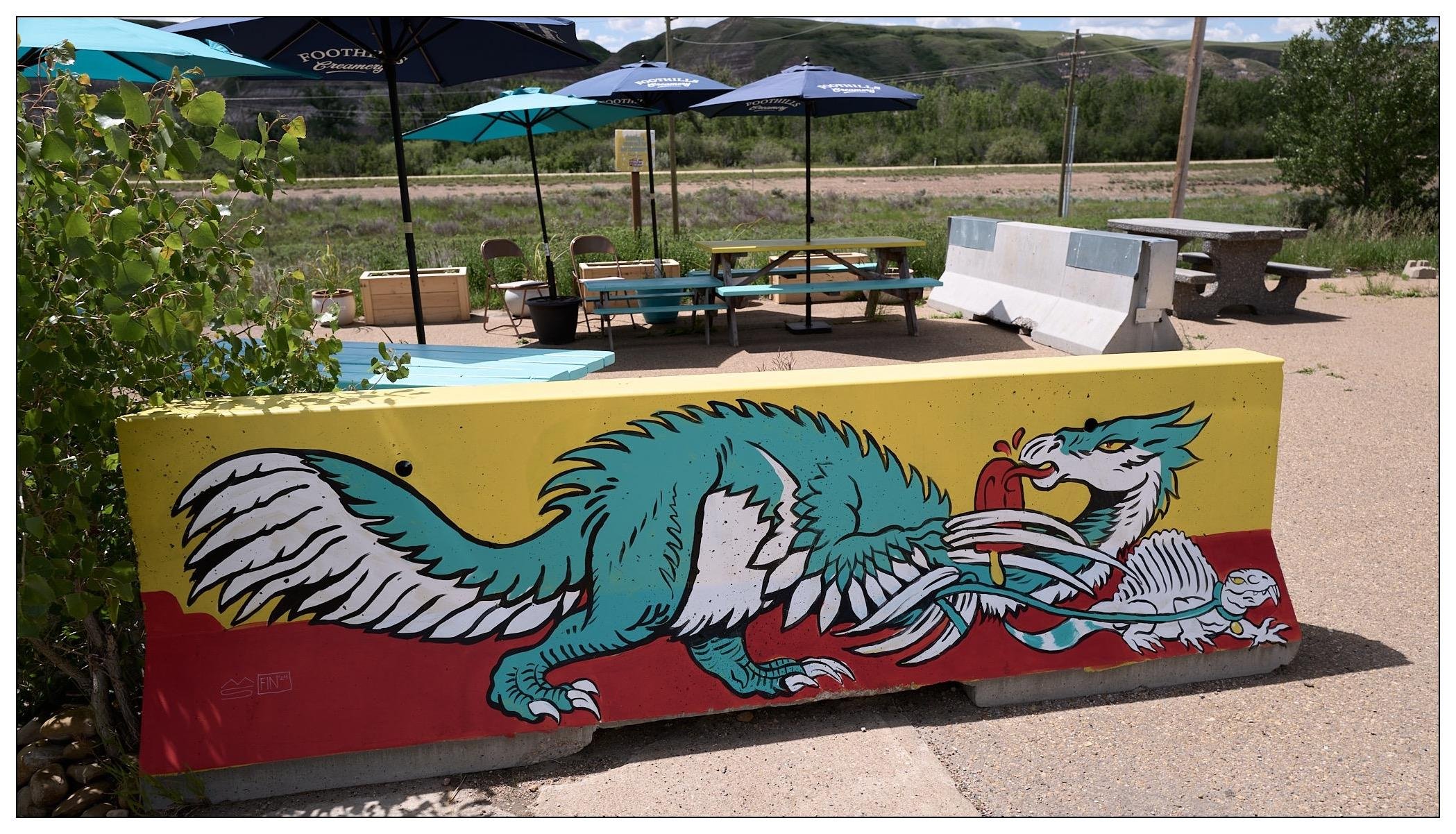 Mural on a cement barrier brick, of a teal and white feathered dinosaur resembling a dragon in some ways.  It is holding and licking a red ice popsicle.   In the background is a seating area with tables and umbrellas.