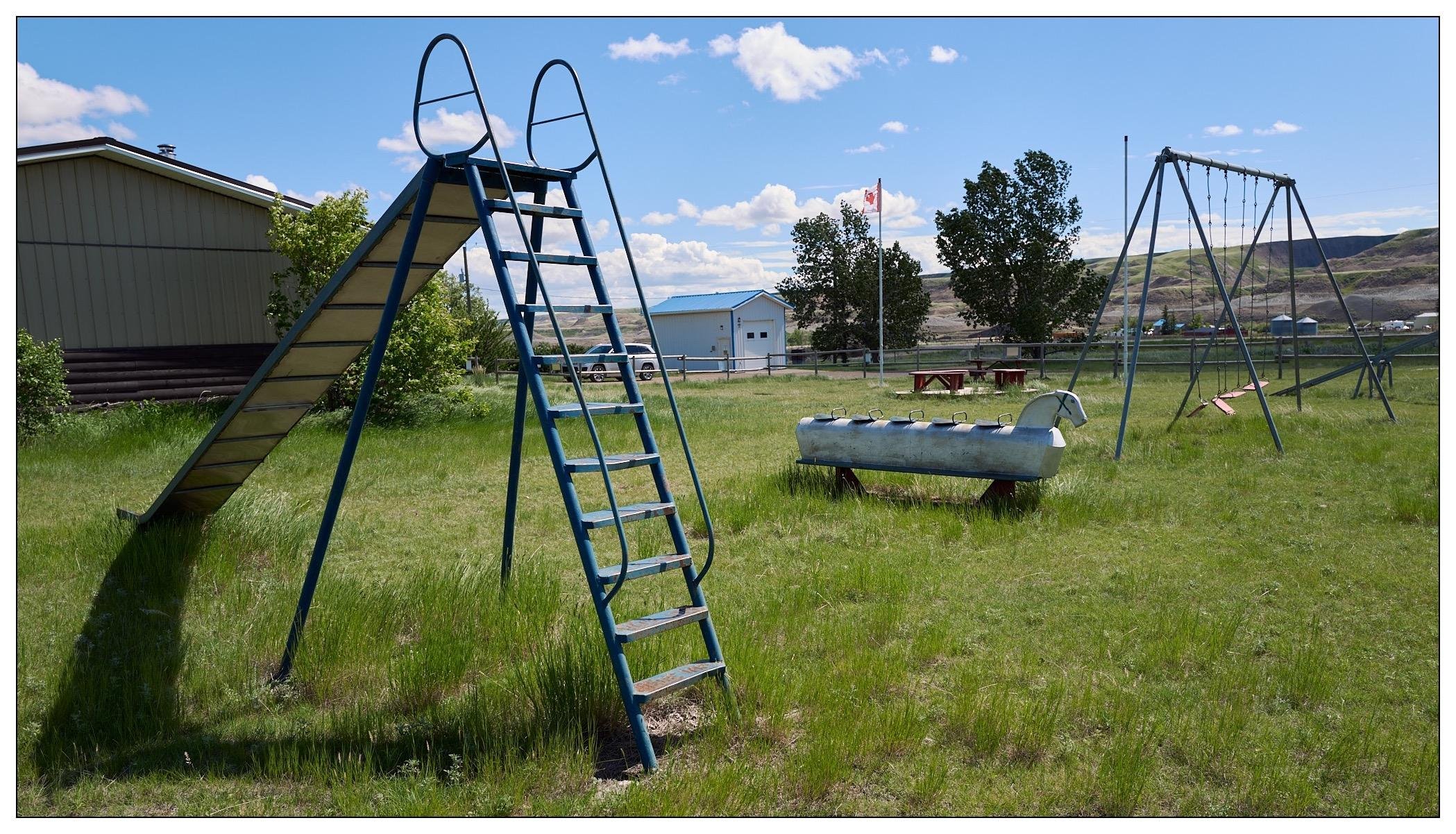 Metal playground equipment in Dorothy, Alberta.  Tall straight metal slide in foreground, then a five seat metal rocking horse, a swing set, and teeter-totter.   It is summer and the grass is very green.