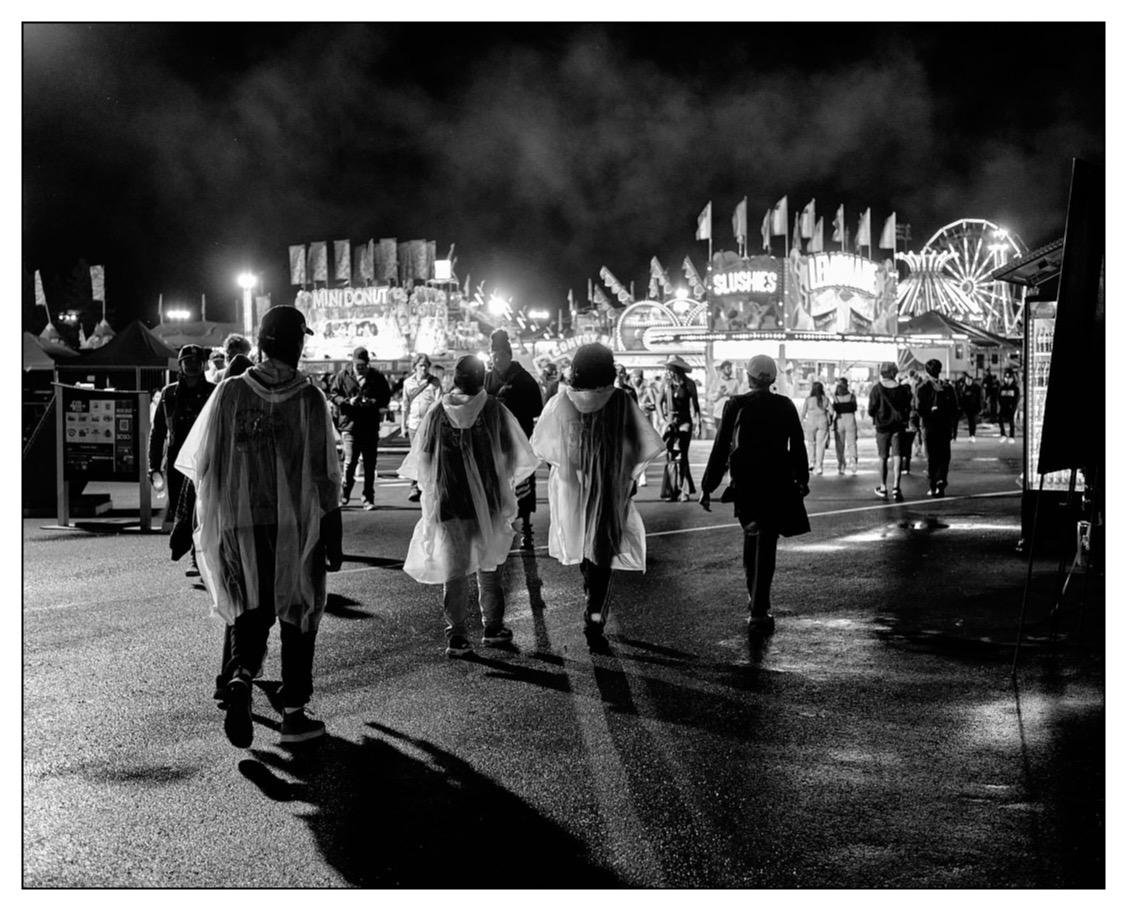 The image depicts a lively night scene at a fair or amusement park. Several people are walking along a wet pavement, casting long shadows due to the lighting around. Some of them are wearing plastic rain ponchos, suggesting it might have recently rained. In the background, there are brightly lit amusement rides, including a Ferris wheel, and various stalls advertising food and drinks like "Mini Donuts" and "Slushies". The mood of the scene is bustling yet casual, captured in black and white, which adds a timeless quality to the photograph.