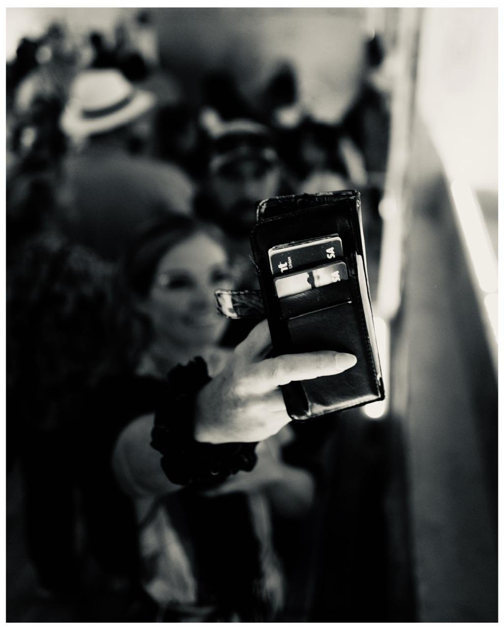 This black and white image depicts a woman holding a smartphone, capturing a selfie or video in a public setting with other people in the background. Her smartphone case has slots with credit cards visible, focusing attention on personal details typically stored within phone cases. The background is slightly blurred, emphasizing the action of the woman and her smartphone. The composition conveys a candid, social moment, likely at a gathering or event.