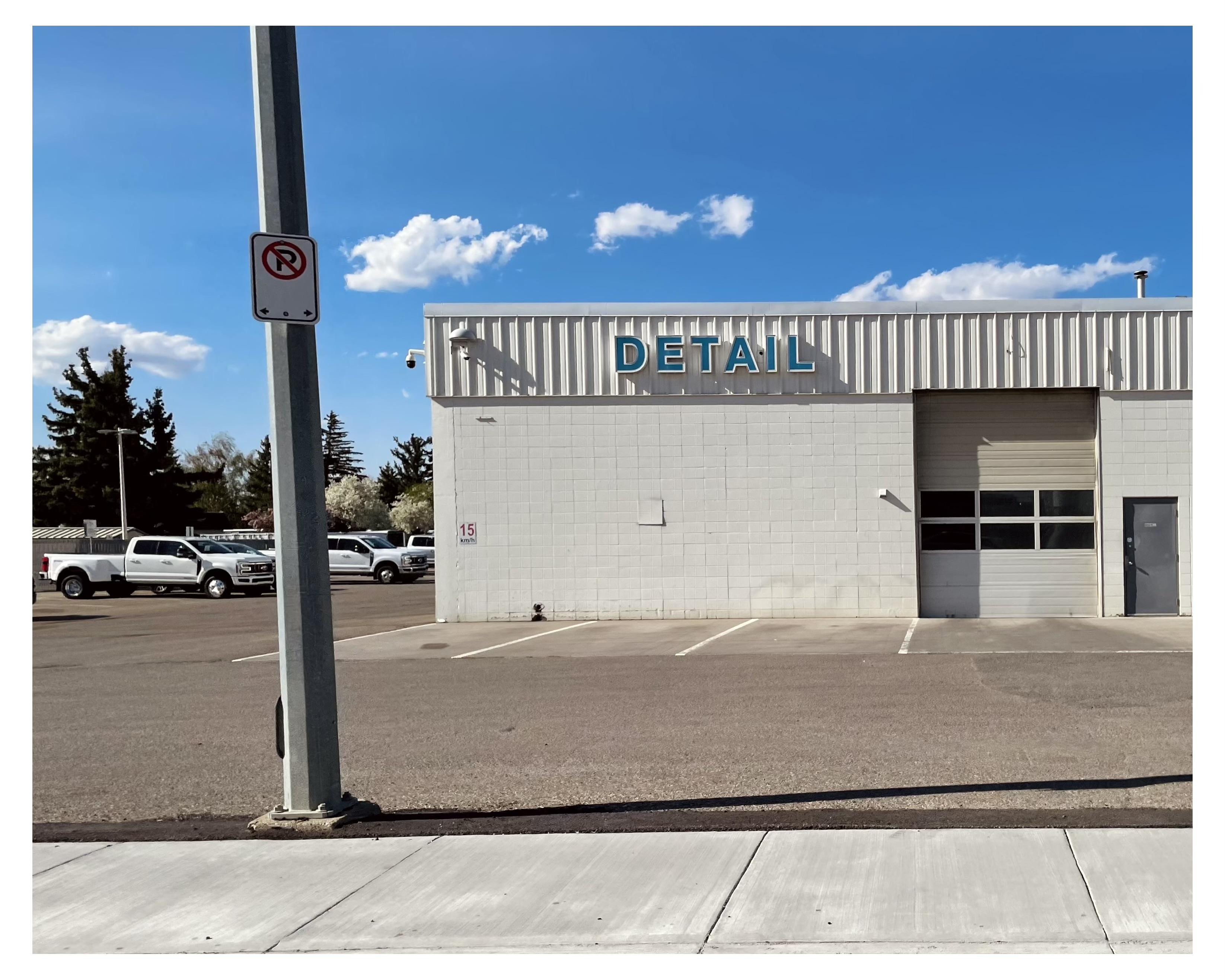 Exterior view of a simple one story white building marked with the word "DETAIL" in large, capital letters on the facade, and a roll up door to the right. 