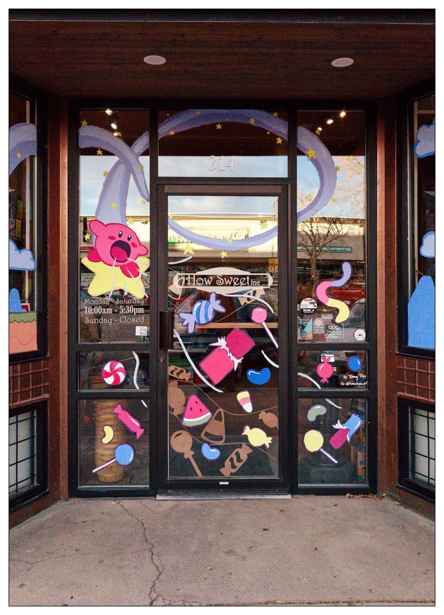 Double glass doors of a candy shop adorned with colorful cutouts of candies and sweets, including lollipops, ice cream cones, and cupcakes. The doors feature a purple banner with yellow stars across the top and are framed by dark wooden panels. The shop's address, 314, and hours are displayed on the doors. Reflective glass shows a bit of the street and other buildings outside.