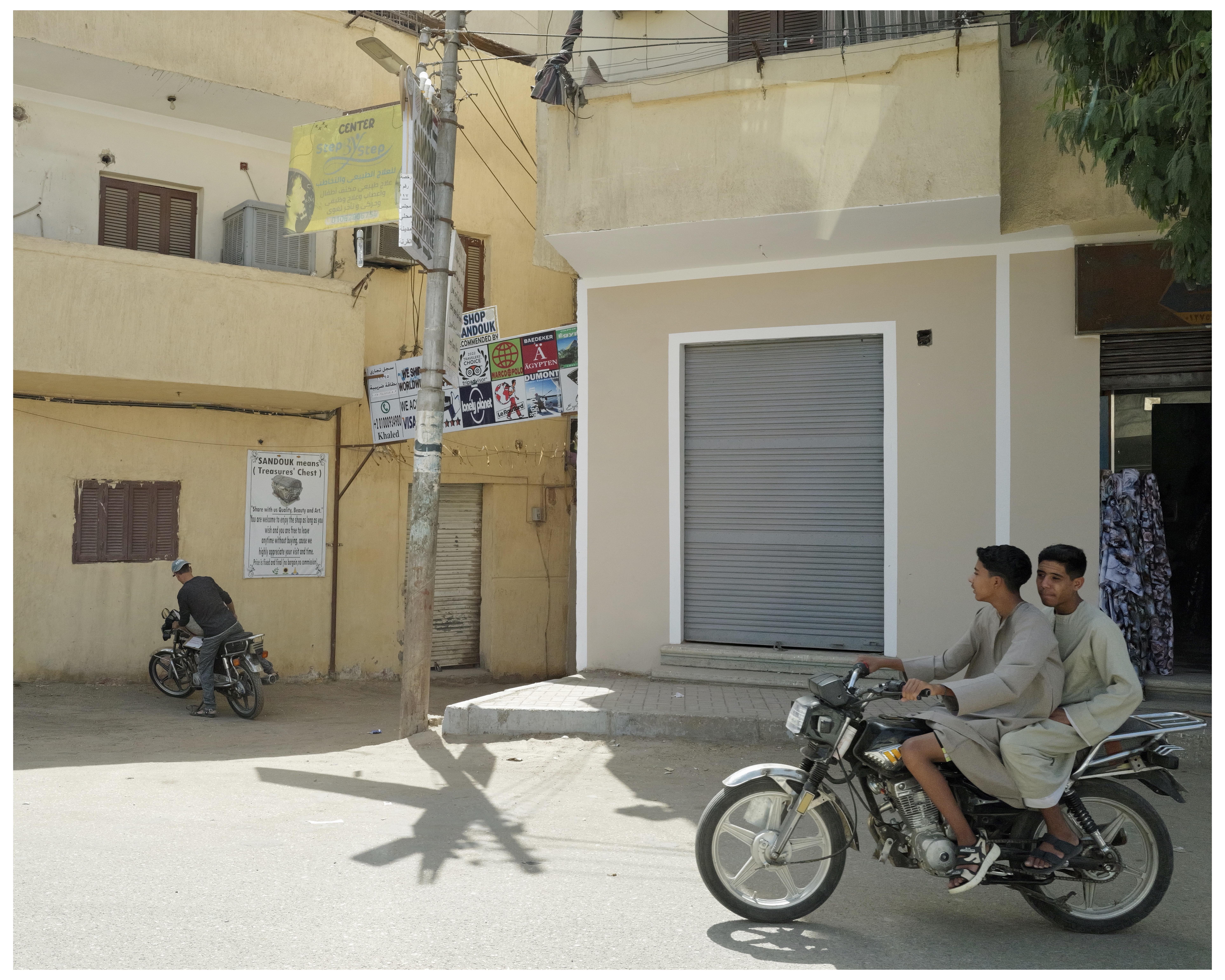 This image depicts a street scene featuring two motorcycles; each is driven by a man with a passenger on one of them. They appear to be in an urban environment characterized by modest buildings, closed shutter storefronts, and advertisements posted on walls and poles. The scene is set under bright, sunny conditions suggesting daylight. The overall atmosphere suggests a routine urban setting possibly in a region where motorcycles are a common form of transportation.