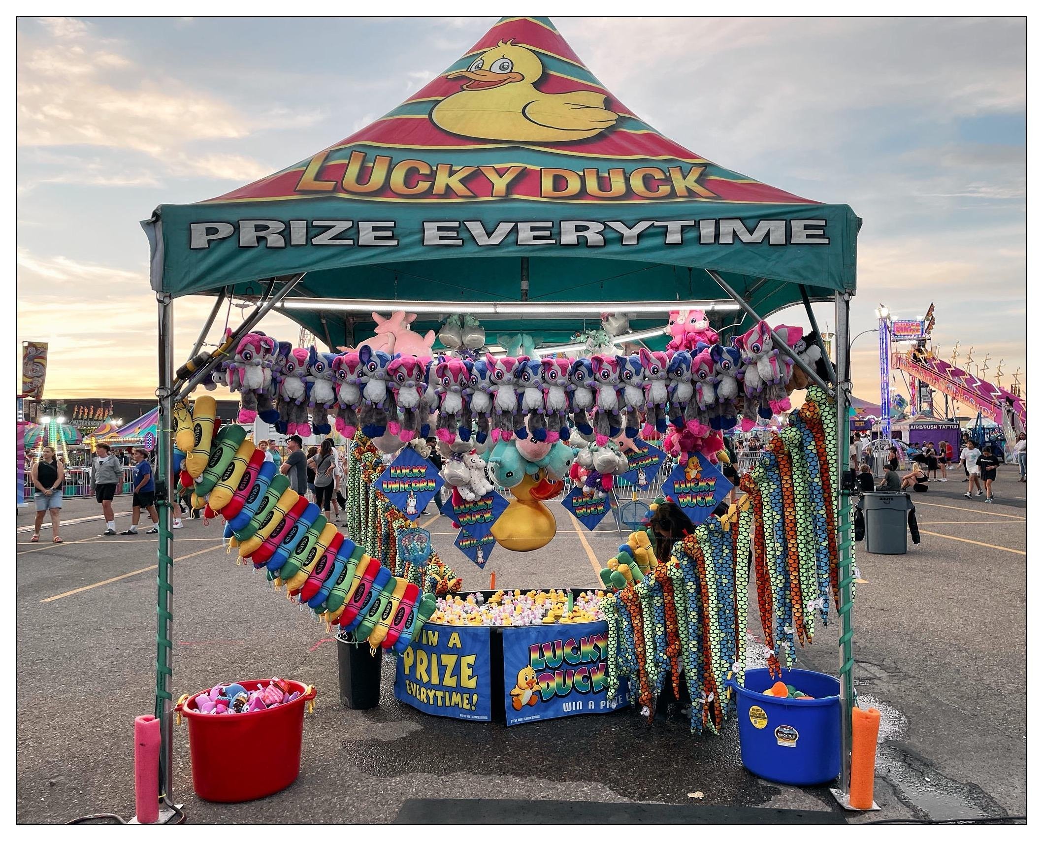 This image shows a colorful game booth at a fair or carnival, named "Lucky Duck Prize Everytime." The booth features various prizes such as stuffed animals (unicorns, ducks) and brightly colored necklaces. There's a pool with yellow rubber ducks for a game, where participants likely pick a duck to win a prize. In the background, you can see a festive environment with other carnival games, rides, and people enjoying the atmosphere. The sky is overcast, suggesting it might be late afternoon or evening.