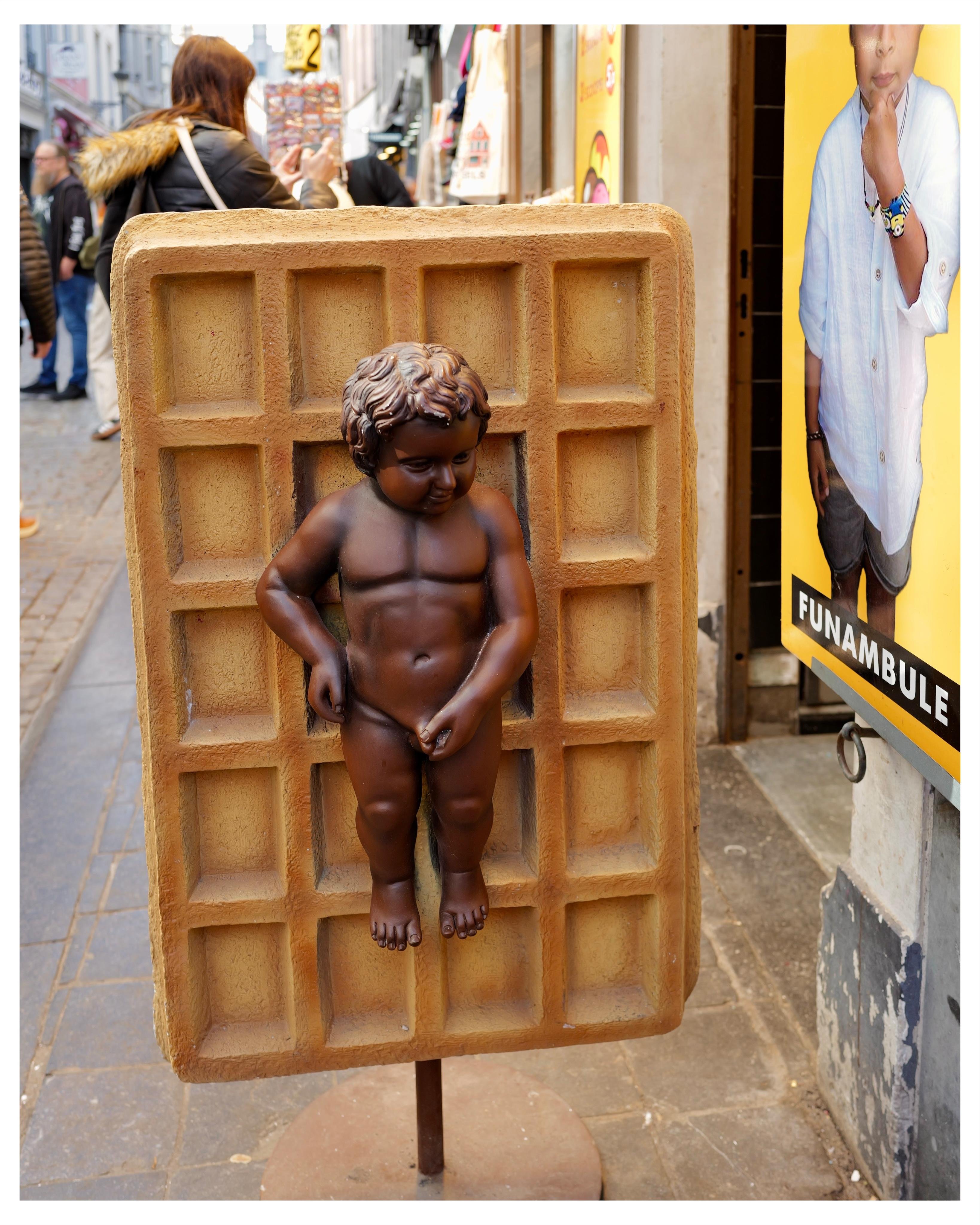 Replica of peeing boy statue (Manneken Pis)as a chocolate effigy on a Belgian waffle.

Advert board on building next to it has a boy rubbing his chin thoughtfully.