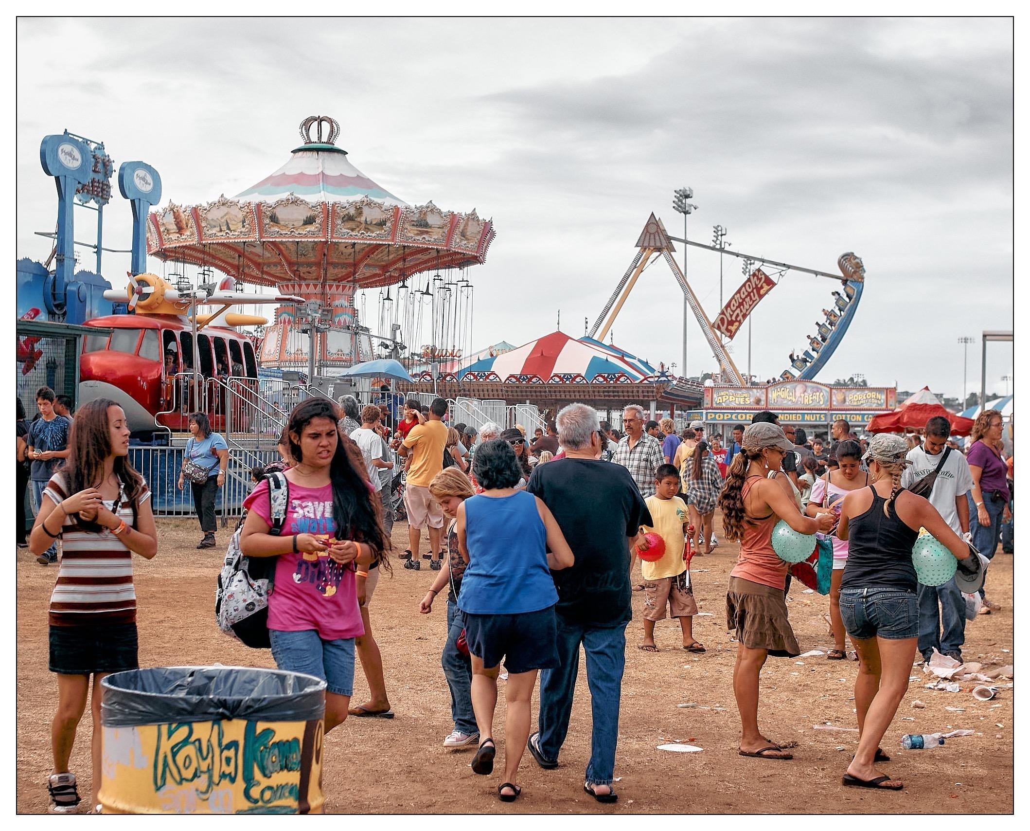 This image depicts a lively scene at a local fair or carnival. There are a variety of amusement rides visible, including a traditional carousel and a large pendulum ride. The area is bustling with people of different ages and backgrounds walking around, enjoying snacks, and interacting with one another. The ground appears to be a dirt surface, indicating the event might be taking place on a temporary setup or in an open field. The sky is slightly overcast, giving the scene a soft, diffuse lighting. Several booths and attractions with colorful decorations and signs such as "Popcorn", "Cotton Candy", and "Mini Donuts" suggest the availability of typical fair concessions. This image captures the casual, festive atmosphere commonly found at such events.