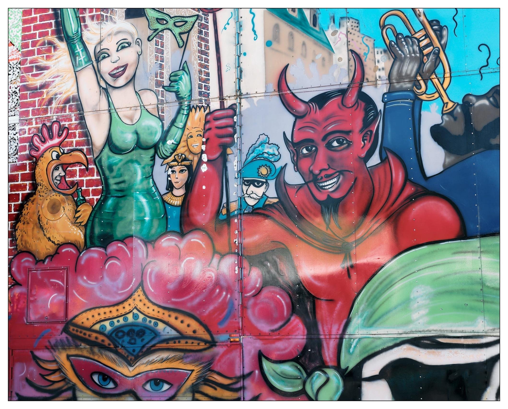 This image features a colorful mural with various whimsical and fantastical characters painted on a wall. On the left side, there is a character depicted as a woman with blonde hair, dressed in a green outfit, raising her arms with one hand holding a mask. Beside her, there is a large red creature resembling a devil with horns, cheerful expression, and muscular physique, holding a golden musical instrument. In the background and between these larger characters, several smaller figures can be seen, including one with blue hair. The artwork is vibrant, utilizing a mix of bright colors.