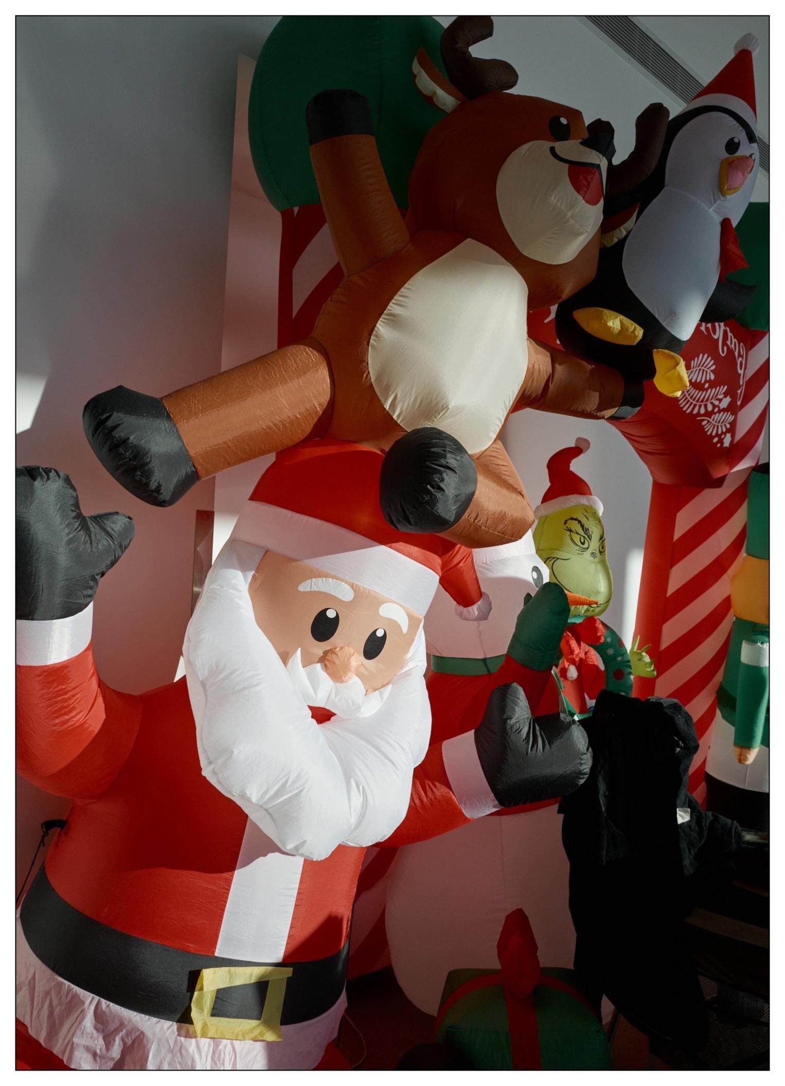 Large inflatable Christmas display featuring Santa Claus in the foreground with candy-cane-striped arms crossed. Behind Santa, several other festive inflatables including a reindeer, a penguin on a polar bear, and the Grinch, each decorated with holiday elements like scarves and hats. The setting appears indoors against a soft-lit wall.