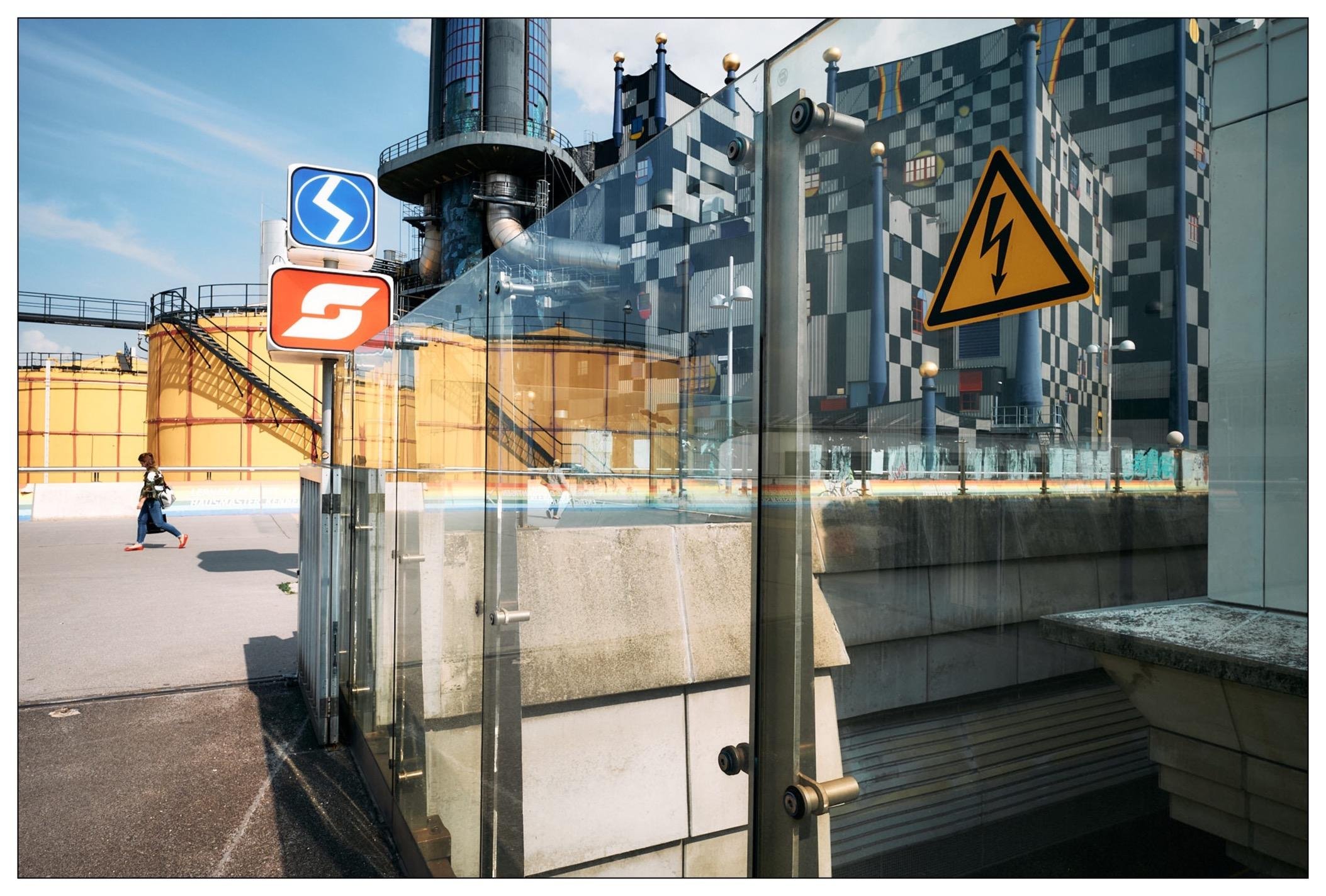 This image features a complex urban scene with multiple reflective surfaces, creating a visually intriguing overlay of elements. This creates a nearly collage-like effect. Specifically, it includes:

1. **Road signs:** There are multiple traffic signs visible including a direction arrow, a speed limit sign, and an electric hazard warning sign.
2. **Industrial Background:** In the background, there appears to be an industrial setting with structures such as a tall chimney stack and yellow scaffolding.
3. **Glass Reflections:** The foreground prominently displays a glass surface reflecting various elements like the road signs, blending them with the actual physical objects behind the glass.
4. **A person:** An individual is visible in the mid-ground, moving through the space on foot, wearing red shoes, adding a sense of everyday human activity to the scene.
5. **Architectural Details:** The photo includes modern architectural elements like sleek glass panels and a stairway, suggesting an urban environment with contemporary design.

Overall, the photo captures a dynamic urban environment where industrial, architectural, and everyday human elements intermingle, highlighted through an artistic use of reflection and perspective.