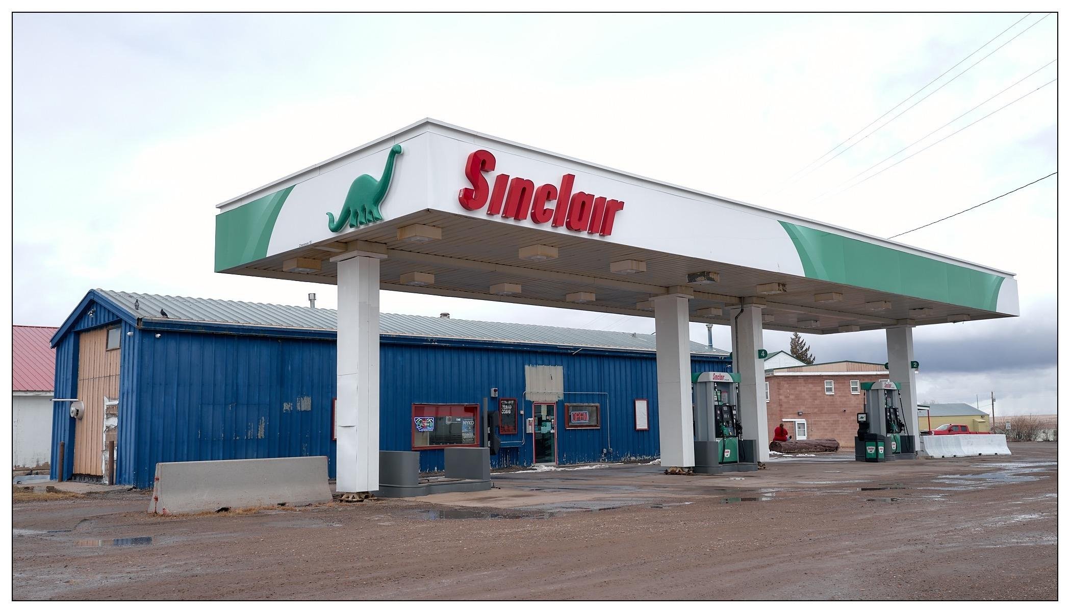 Sinclair gas station. The station features several fuel pumps under a large canopy with the Sinclair logo and their distinctive green dinosaur mascot. Adjacent to the canopy, there's a blue building that could be the station's convenience store or service area. The sky is overcast and the ground appears wet, suggesting recent rain or snow. The setting seems somewhat rural or semi-rural, given the sparse surroundings visible in the background.
