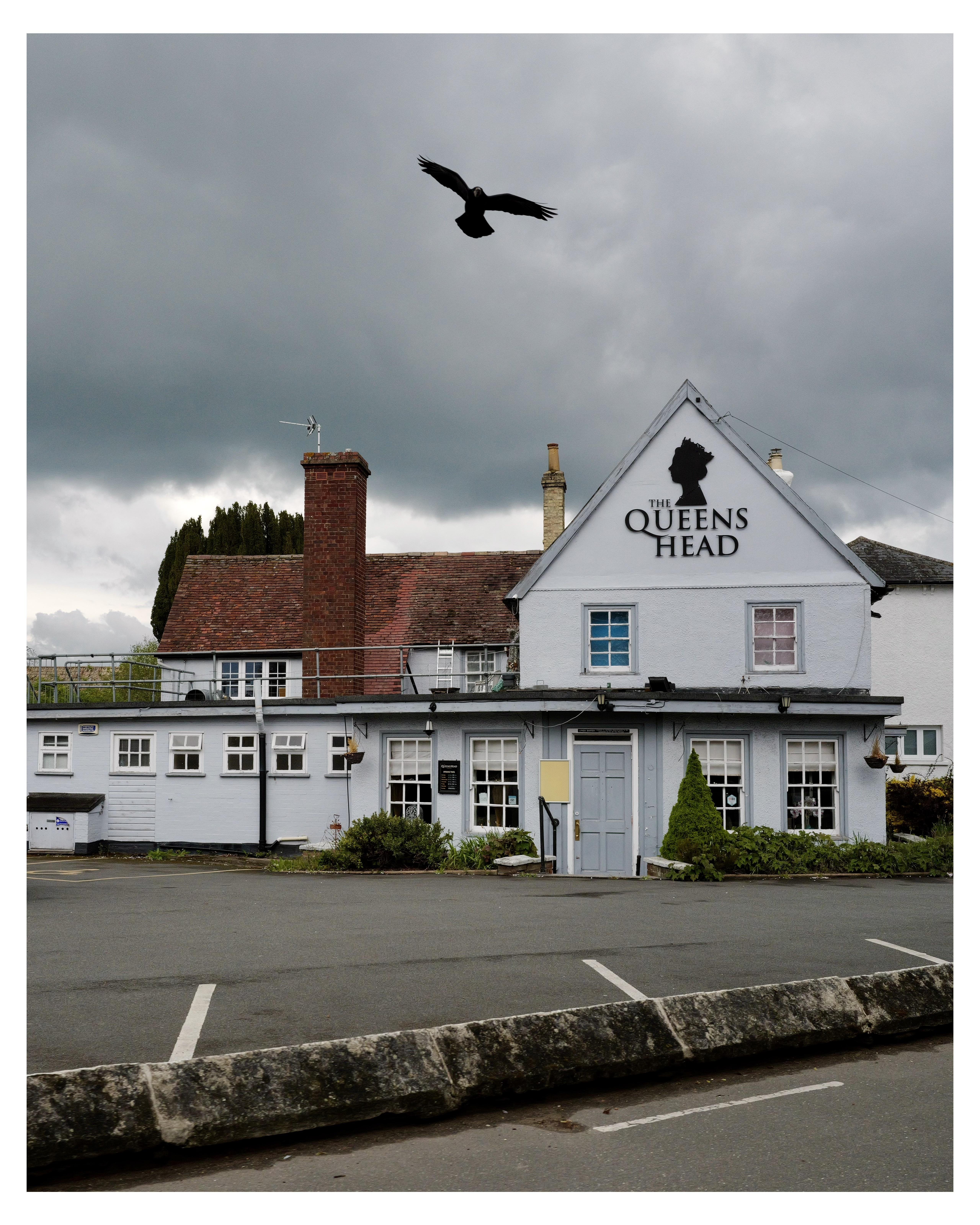 The image shows a two-story building called "The Queens Head," which appears to be a pub, given the traditional architectural style and signage. It has a clean white facade with blue trims around the windows and doors. The building is contrasted by a gloomy, cloudy sky, which adds a dramatic atmosphere. A bird, possibly a crow or raven, is flying in the sky above the building, enhancing the moody feel of the scene.