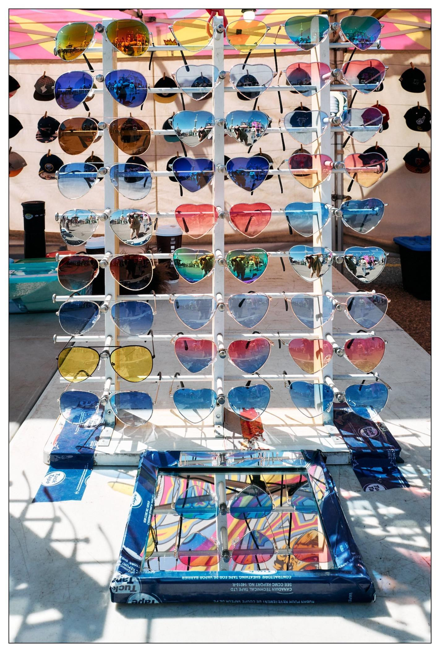 Vertical display racks holding various sunglasses with reflective and colored lenses including multiple heart-shaped frames. Colors range from vibrant pinks and blues to yellow and mirrored reflections. Sunglasses stand against a light background, under sunlight that casts shadows on the table below, which features a mirror that is tuck taped to the table.
