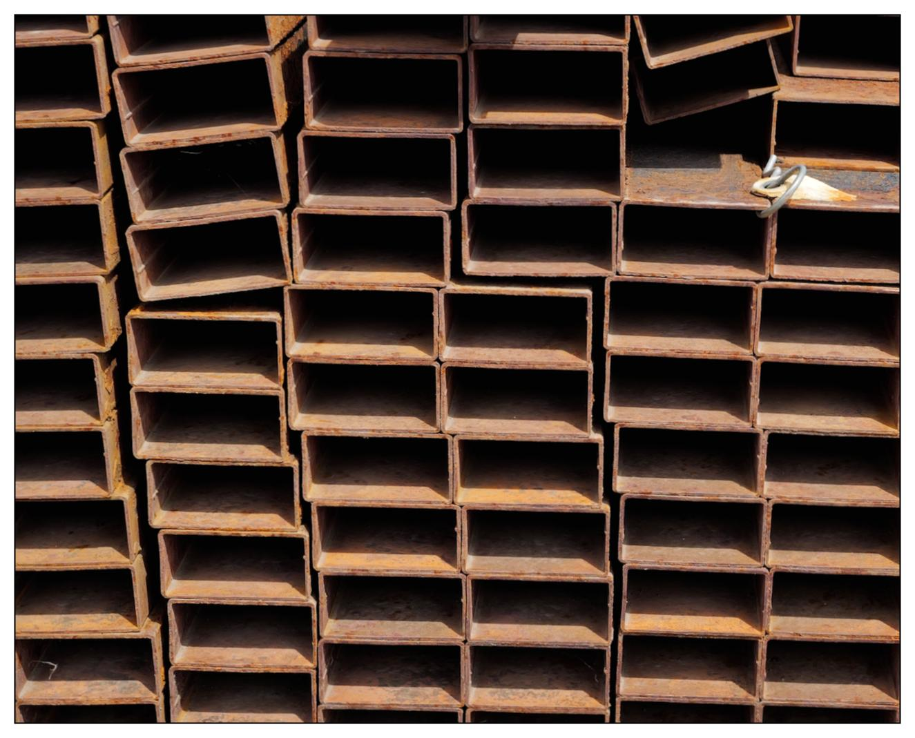 Stacked rusty metal channels form a grid pattern with square and rectangular hollow sections. Some compartments are slightly bent or distorted, and a small piece of debris rests on one horizontal section towards the right.