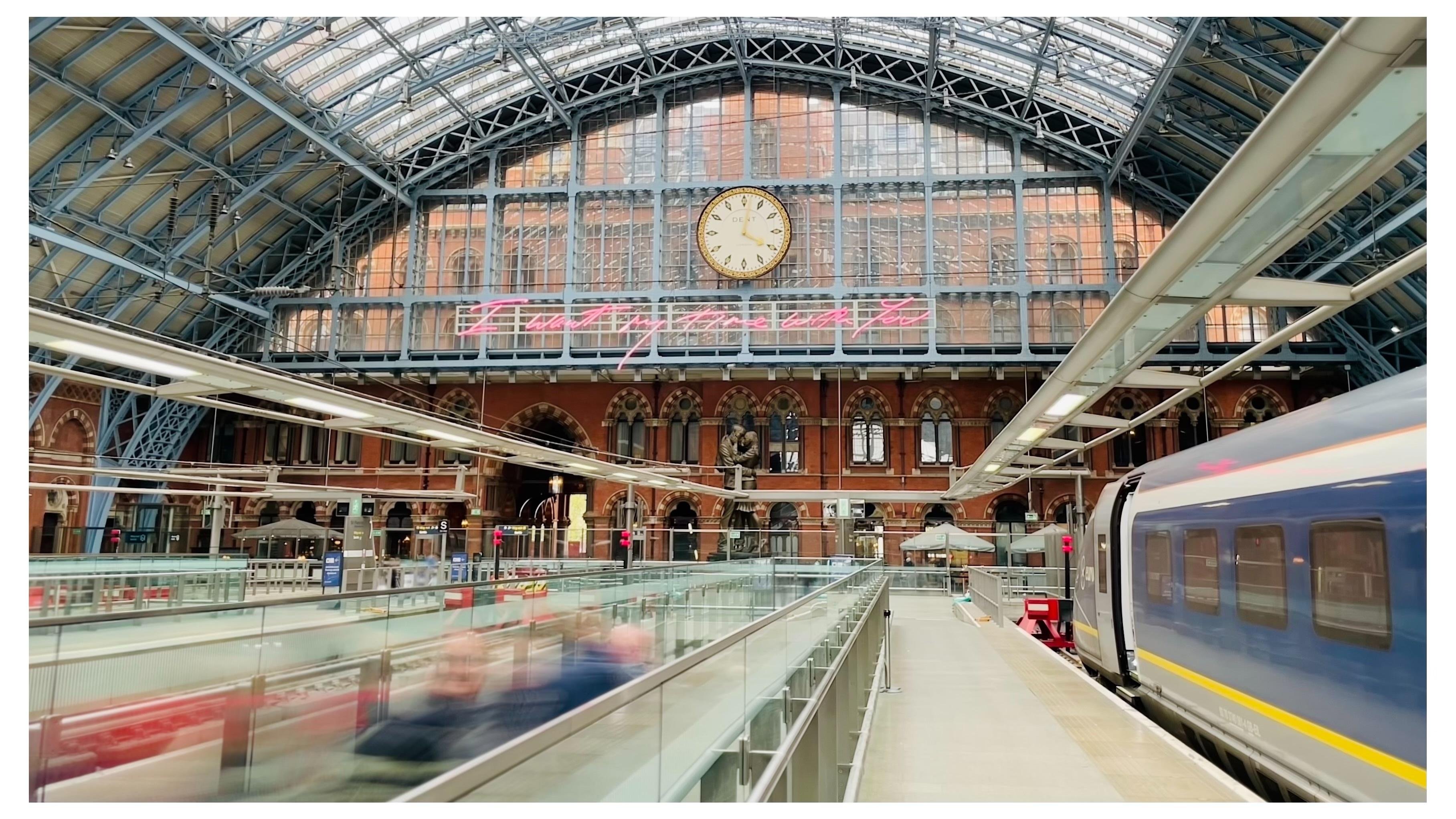 Subject motion blur, people moving down a sliding sidewalk at a train station.  Glass canopy overhead, Eurostar train on right.