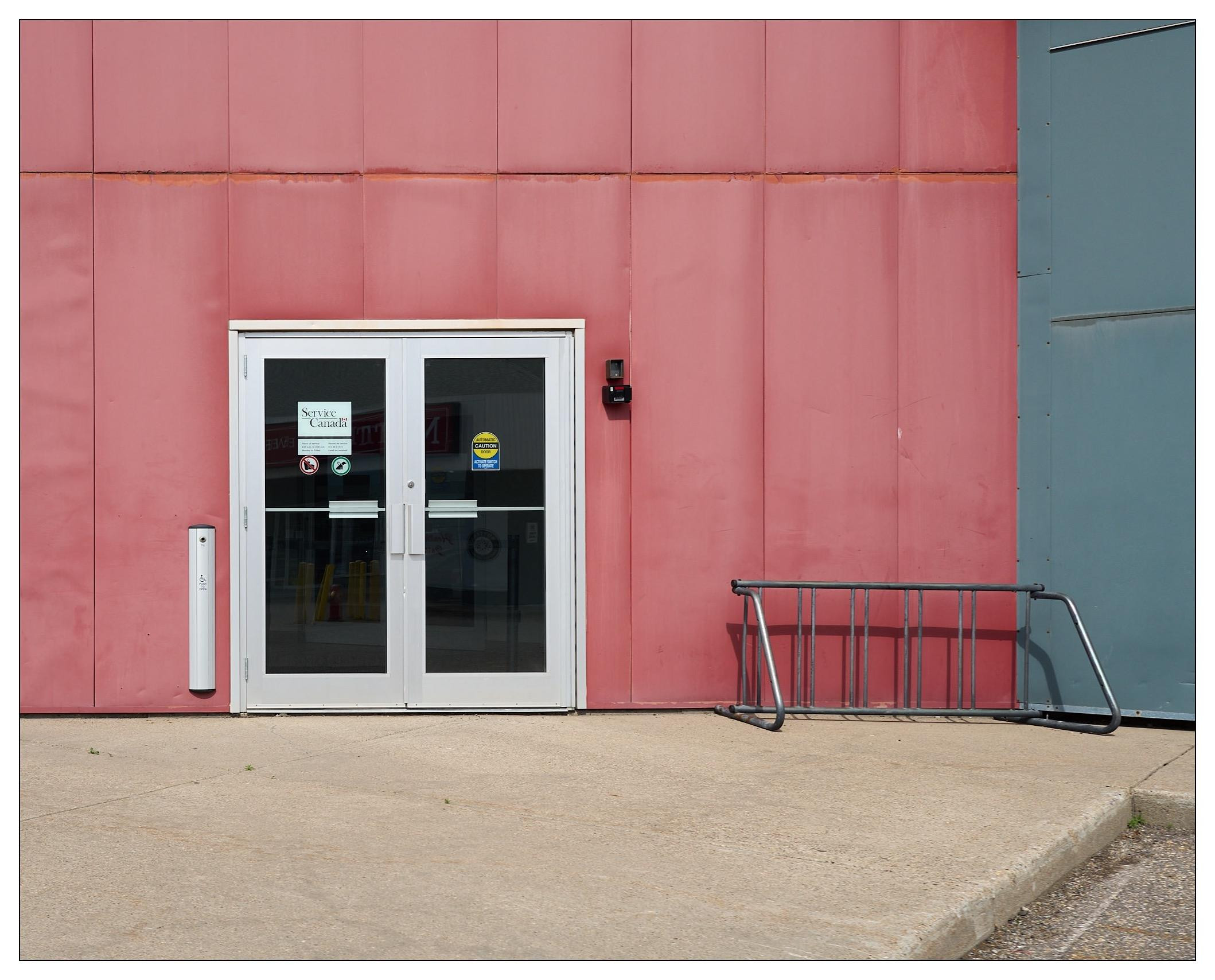 Double doors with white frames set in a wall divided into red and bluish-gray panels, with signs including "Notice Security Cameras in Use" posted on the glass. The doors are flanked on the right by an empty, tipped metal bike rack on a concrete ground.