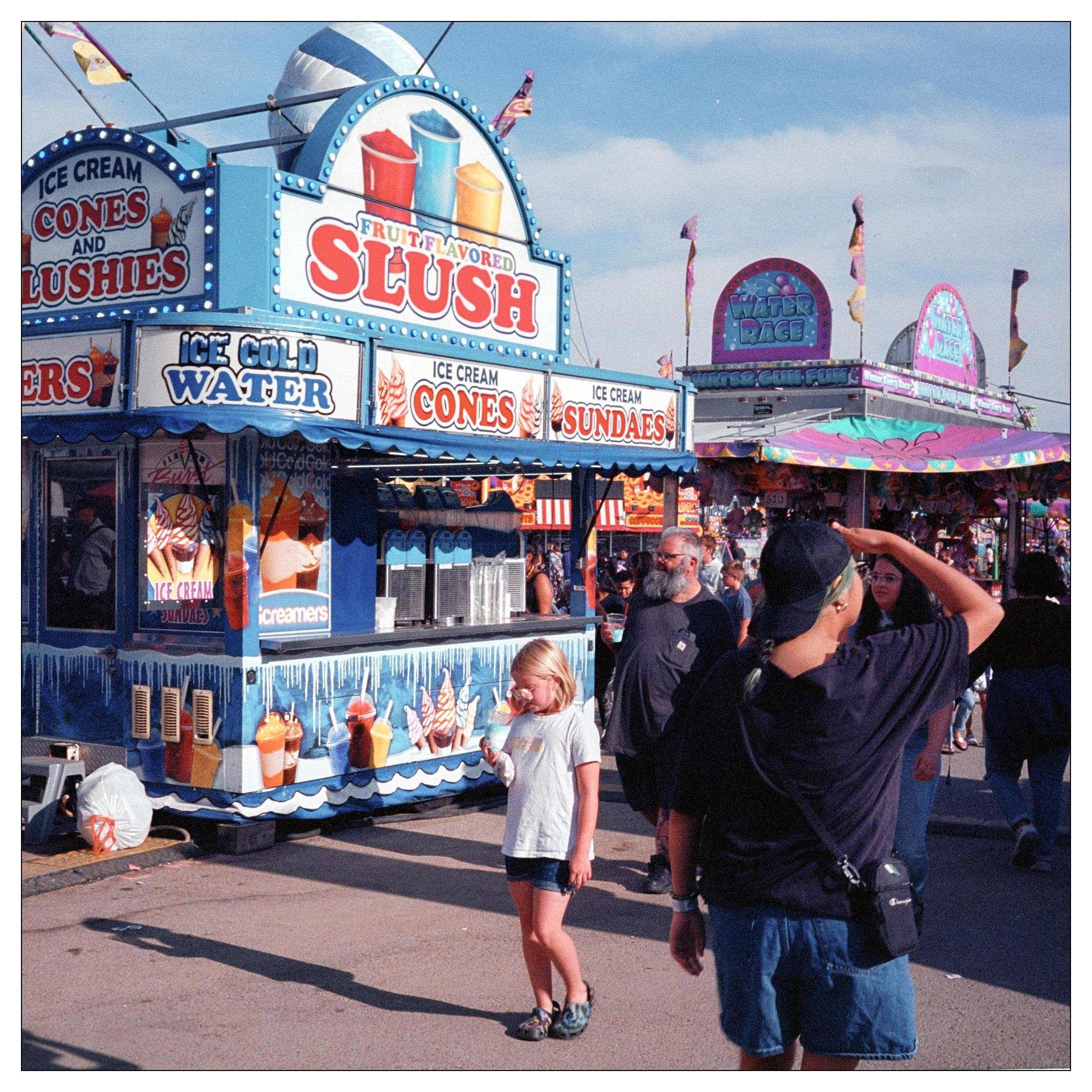 This image captures a lively scene at a fair or outdoor festival. It features a colorful food stand advertising "Fruit Flavored Slush," "Ice Cream Cones," "Sundaes," and "Ice Cold Water." The stand is decorated in vibrant blues and yellows, and it's bustling with activity around it.

There are several people in the image, including a young girl standing in the foreground, almost center frame, who appears to be eating or holding something close to her face. To her left, an adult male is seen from behind, wearing a black T-shirt and a camera around his neck, and he's shading his eyes with his hand.   The triangular space created by his arm and head is occupied by the face of a woman walking towards them.

The overall atmosphere suggested by the image is vibrant and festive, typical of a fair with food stands and possibly rides, as hinted by more structures in the background that resemble part of an amusement ride setup. The time of day seems to be during daylight hours, under clear skies, contributing to the bright and cheerful setting.