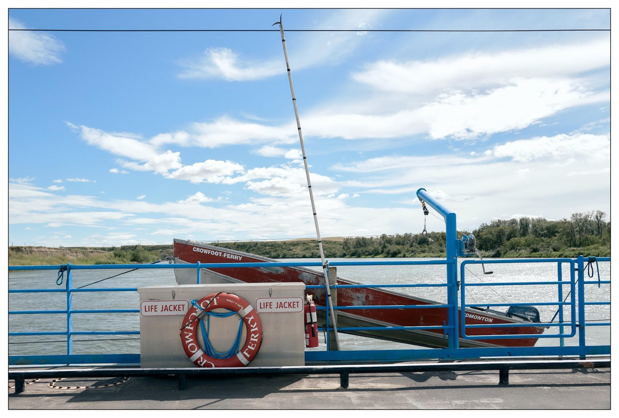 A ferry service boat with a bright red and blue color scheme is stored on the rail of the ferry itself. The ferry rail is painted blue, with a life preserver attached to it, set against a backdrop of a calm river under a blue sky with scattered clouds. A green, tree-lined riverbank is visible in the distance.