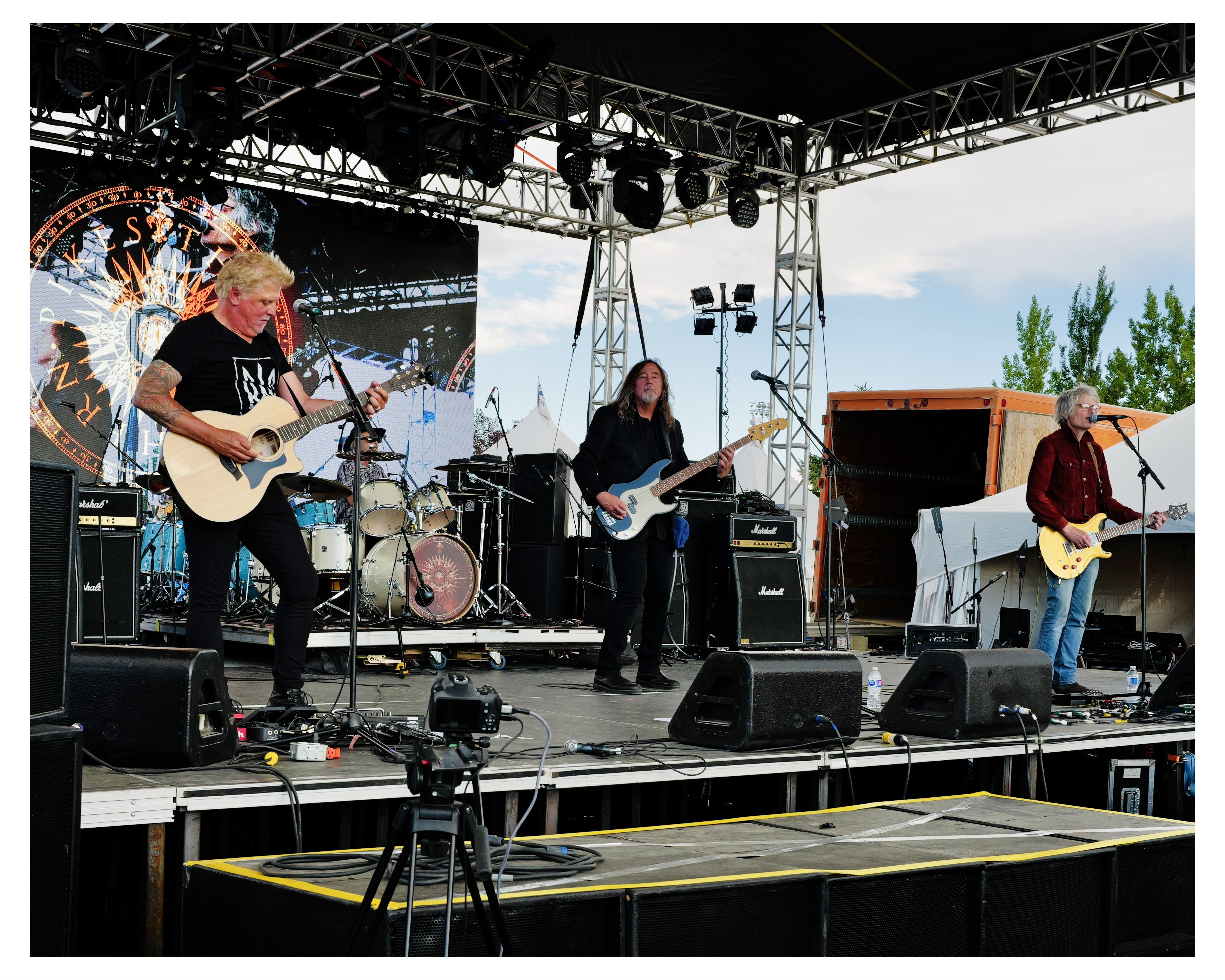 Four band members perform onstage at an outdoor music concert. On the left, a man with platinum blonde hair plays an acoustic guitar. Next to him, a long-haired drummer sits behind a drum set, and a bass player with black hair stands playing a blue electric bass. On the far right, another guitarist with gray hair plays a yellow electric guitar. The stage is equipped with large Marshall amplifiers and several microphones. A large banner with ornate designs and the word "STARZ" hangs in the background, supported by a metal truss structure with stage lights.