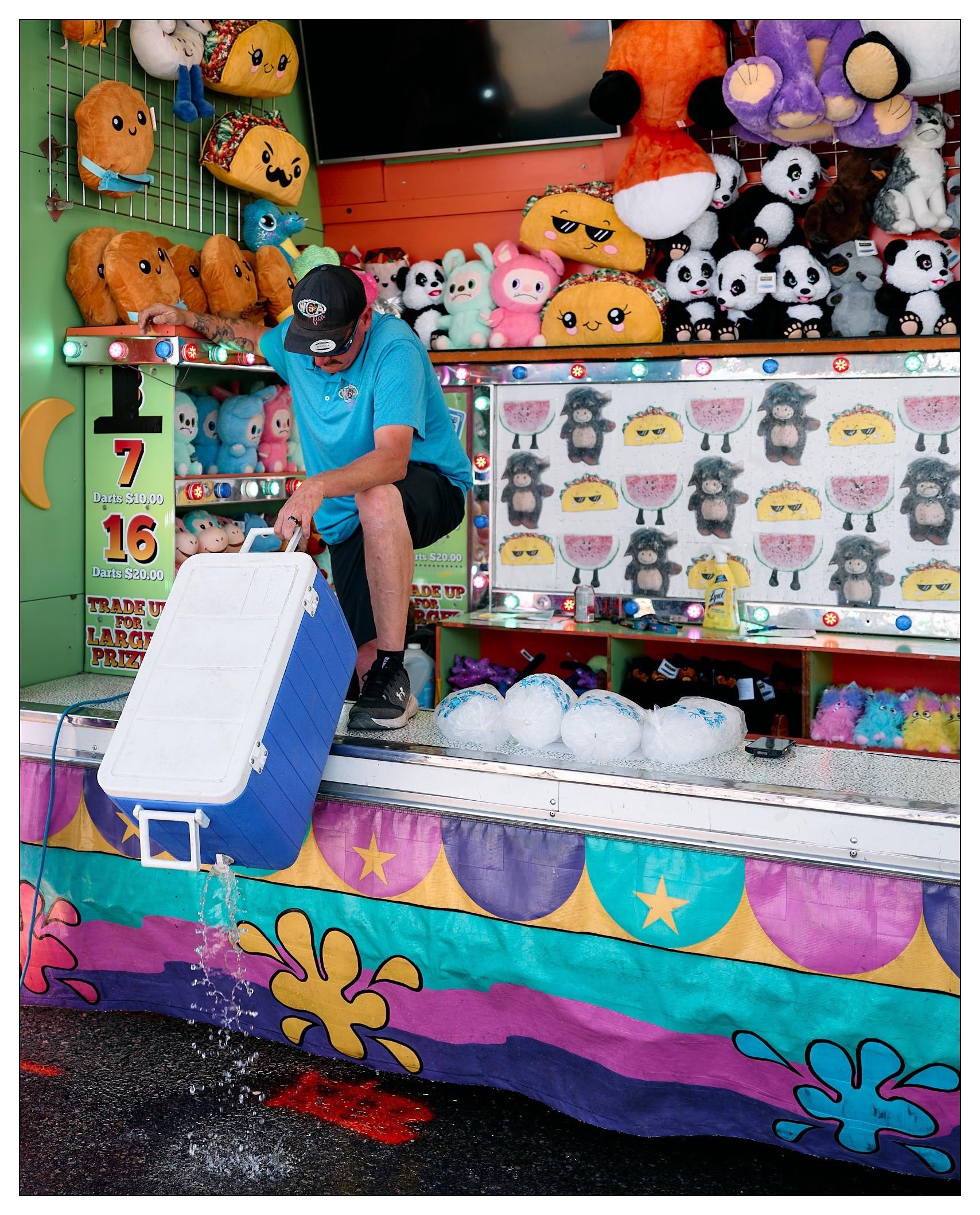 Man in a blue shirt and cap, seated at a carnival game stall, emptying water from a cooler. The stall is decorated with colorful artwork and arrayed with various plush toys like emojis and pandas as prizes. Visible game targets feature cartoon sheep faces, and the booth has a vibrant, multicolored base with star and flower motifs.