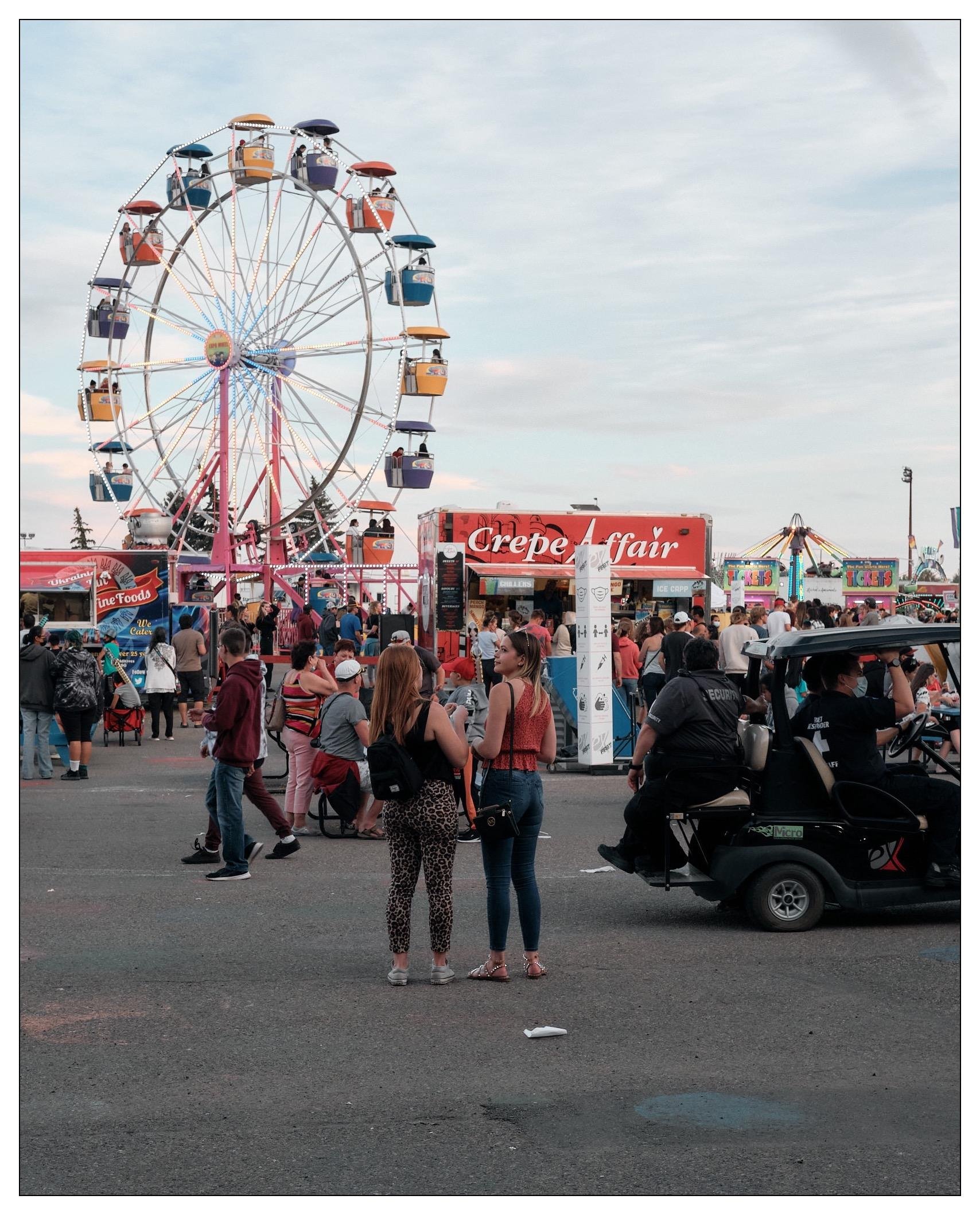 The image shows a lively carnival scene featuring a large Ferris wheel with colorful gondolas against a cloudy sky. In the foreground are people strolling and socializing in an area surrounded by various food stalls, including one named "Crepe Fair." There are two women in the center facing towards each other and talking; others are walking or gathering around. Additionally, several carnival rides and game booths can be seen in the background, creating a festive and vibrant atmosphere typical of a fairground.