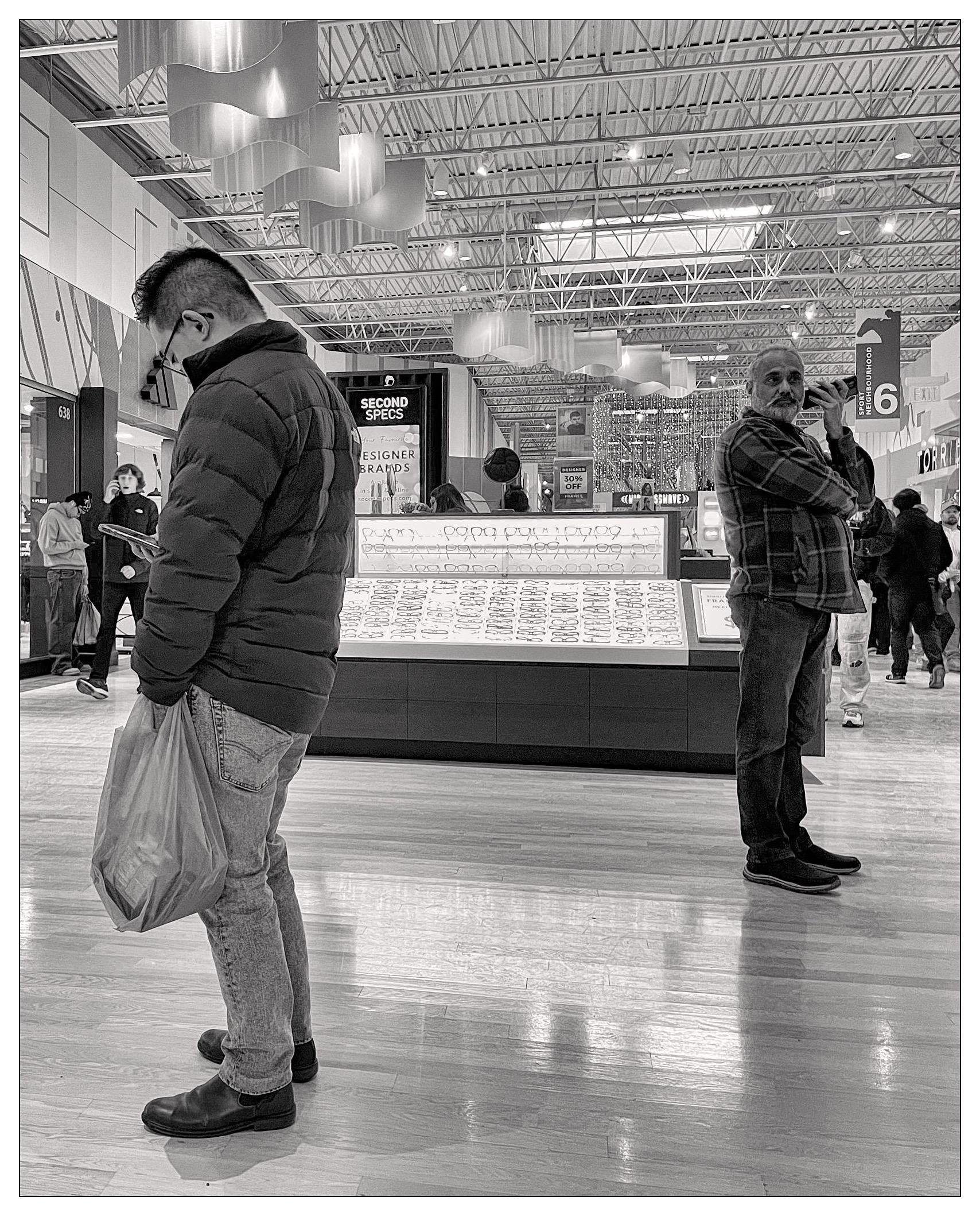 Several people, not together, some foreground and some background, all using their mobile telephones while standing or walking in a shopping mall.   Two are getting very bad neck strains from peering downwards into the screens.   

One is holding his flat with the bottom end to his ear (incorrectly using speaker phone in public), and one actually knows how to talk on a phone call and hold it to his head correctly.