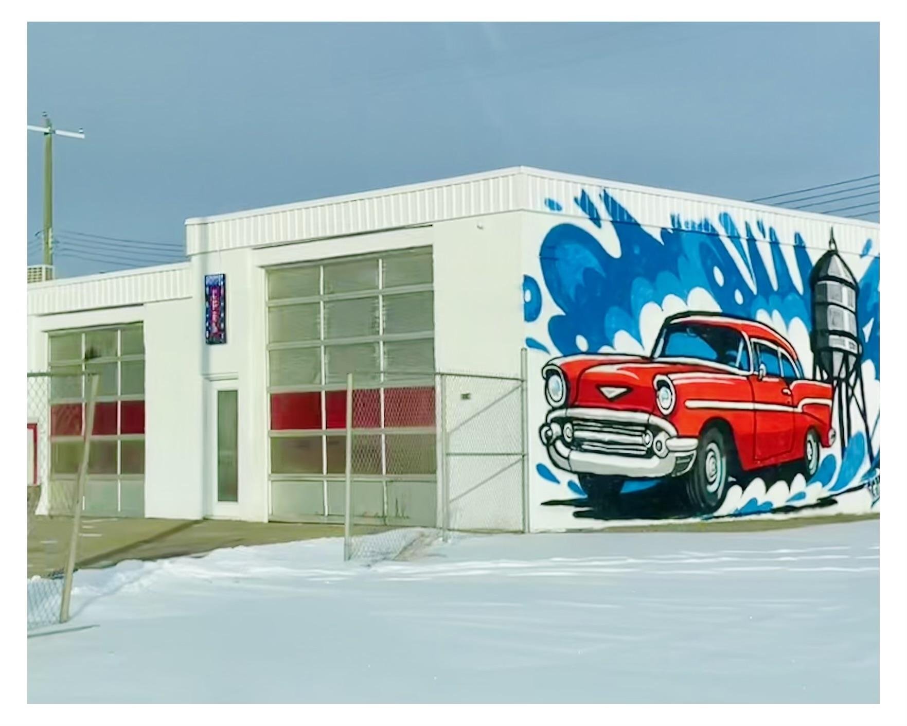 A mural of a red vintage car against a vibrant blue background with white splashes decorates the wall of a white, single-story building. The building, partially surrounded by a chain-link fence, features a glass garage door and several windows with red accents. Snow covers the ground, reflecting the sunny but cold atmosphere.
