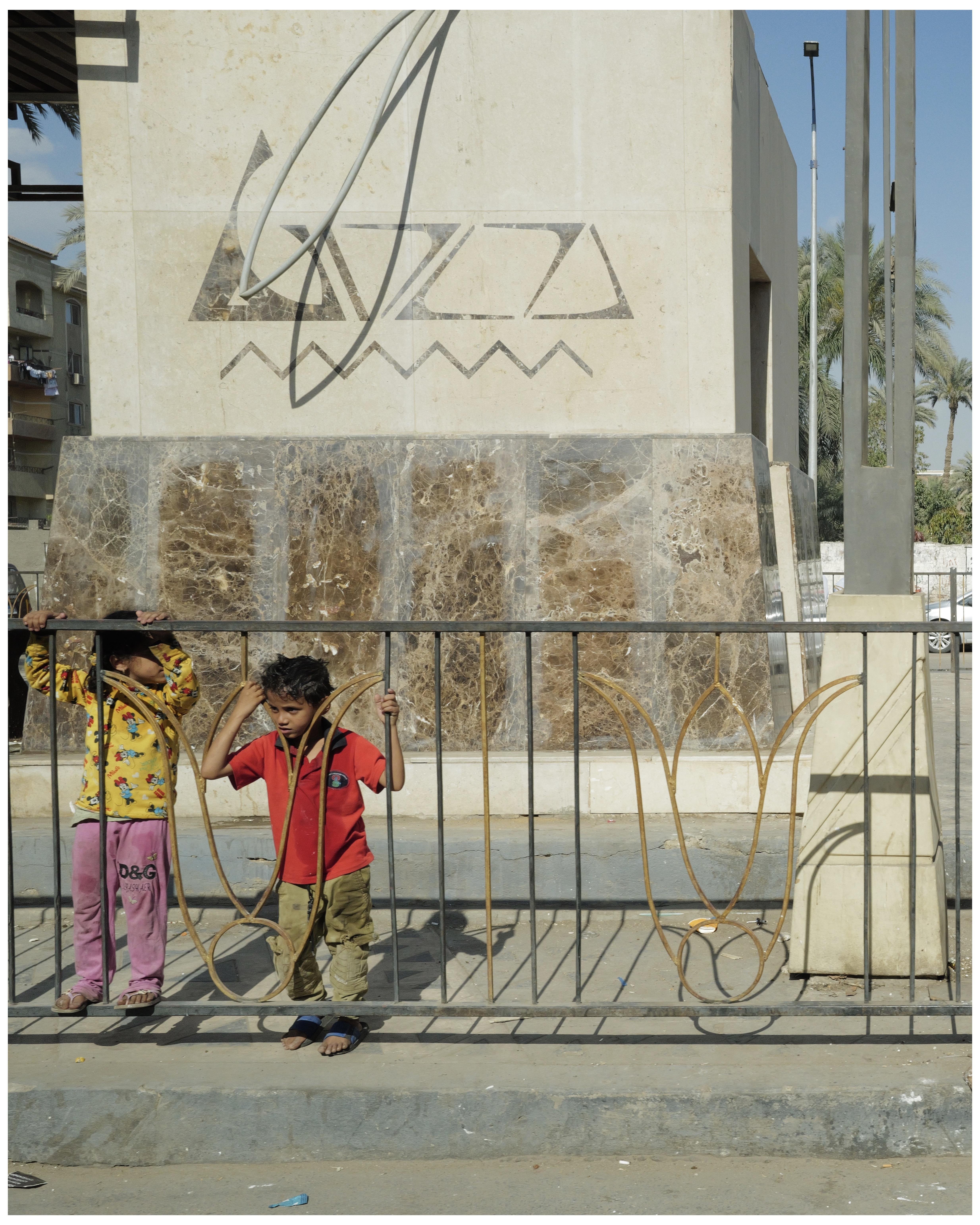 The image shows two children standing by a decorative metal fence with a large, textured stone structure in the background. The structure has a graphic design etched or painted on it that includes abstract geometric shapes and lines. The scenery appears to be in an urban area as there are buildings and a street lamp visible in the background. The children are wearing casual clothing; one child is in a yellow patterned top and pink pants, the other in a red top and green pants. The setting seems to be sunny, likely outdoors in a public or communal space.