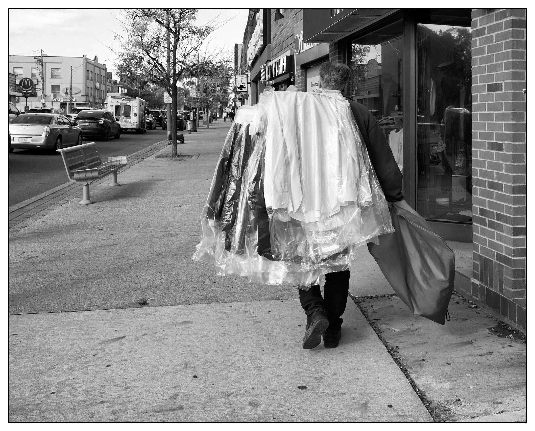 Black and white photograph of a person walking on a city sidewalk, their back to the viewer, carrying a large bundle of dry cleaned clothes covered in transparent plastic. The street is lined with shops and parked cars, with a metal bench and a leafless tree visible to the left side of the frame.