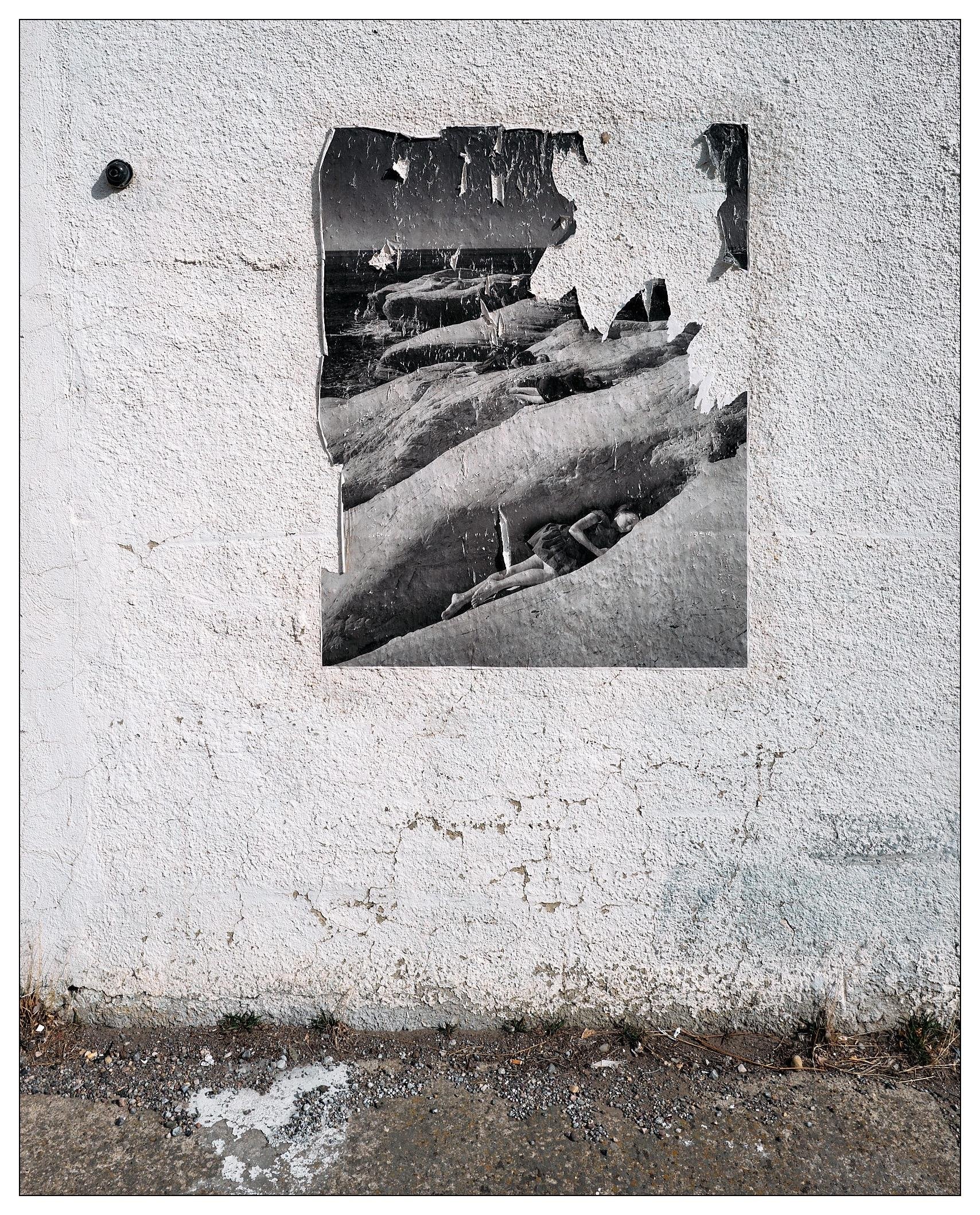 The image shows a weathered poster adhered to a textured white wall. The poster, which is partially torn and peeling, depicts what appears to be a historic, possibly black-and-white photograph of people, likely men, in small boats. Below the wall, there is a strip of ground with some dirt and small stones. The photo generally conveys a sense of decay and abandonment.